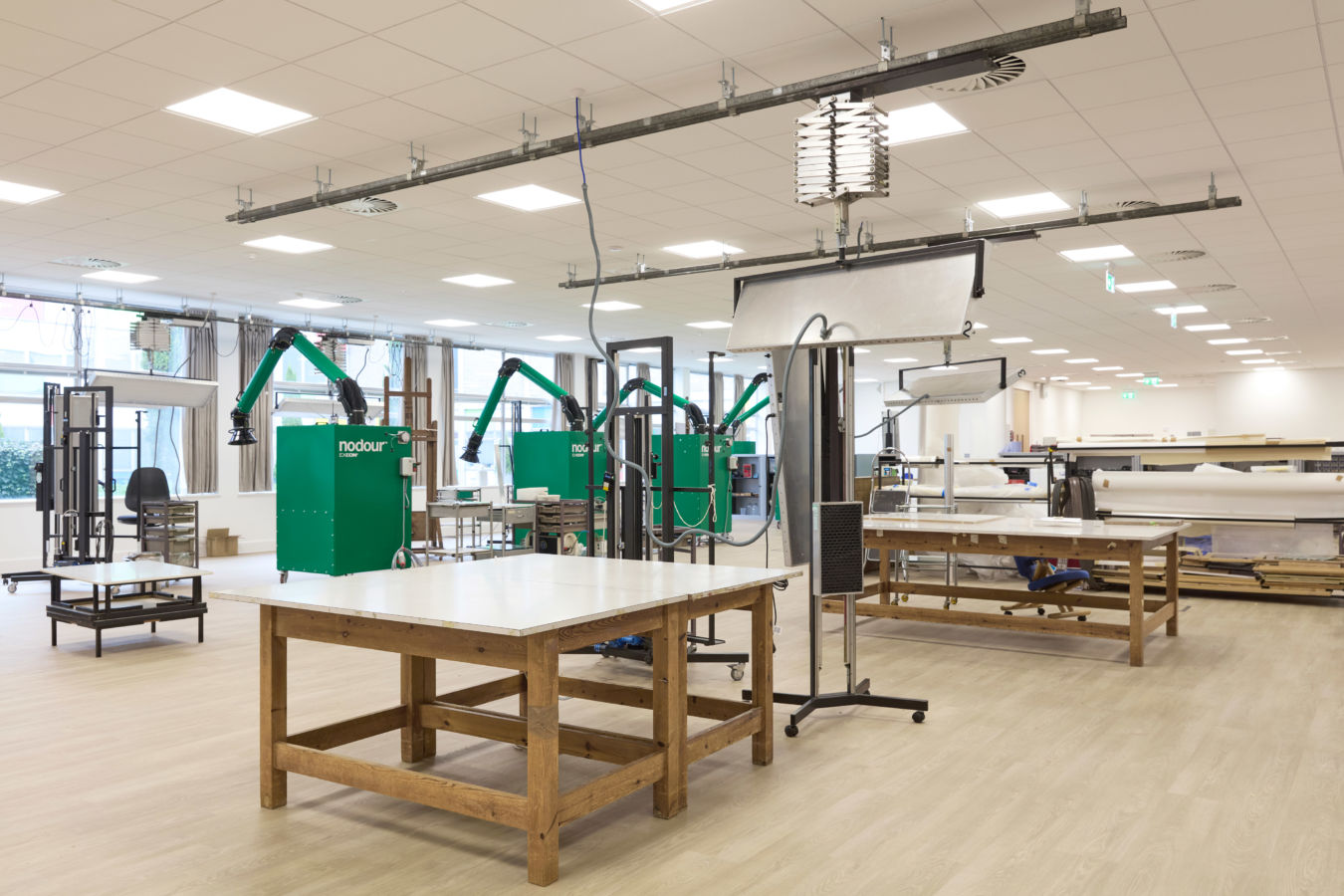 Spacious workshop with wooden tables features green ventilation units labeled "nodour" and extendable hoses, set in a bright room with white ceiling panels and large windows.