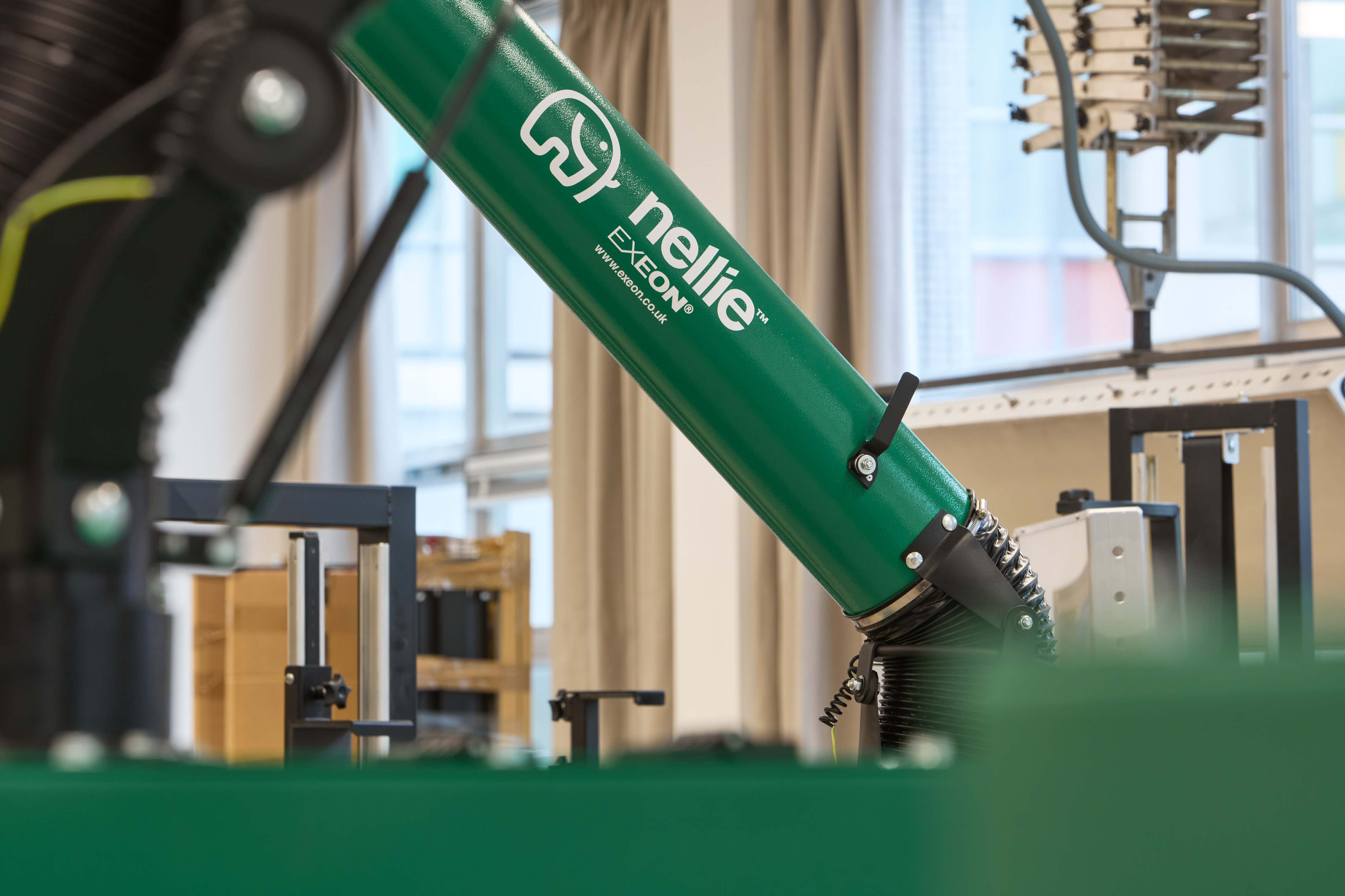 Green industrial equipment labeled "nelie," "EXEON," and "www.exeon.co.uk" is positioned diagonally in a workshop with metal structures and windows in the background.