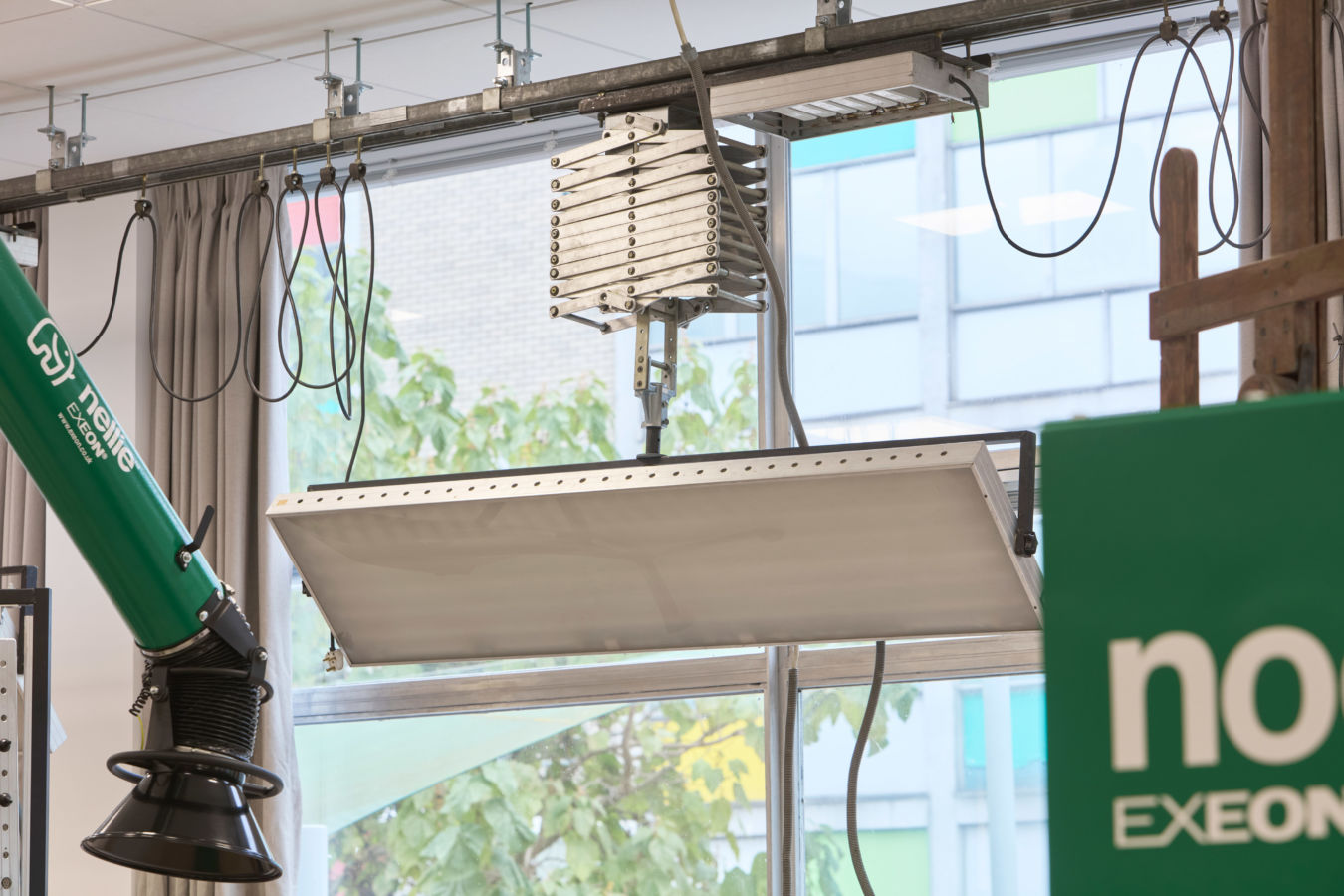 A metal ventilation hood attached to the ceiling, surrounded by hanging cables, sits next to a green "Nederman Exeon" extractor in a room with large windows and trees outside.