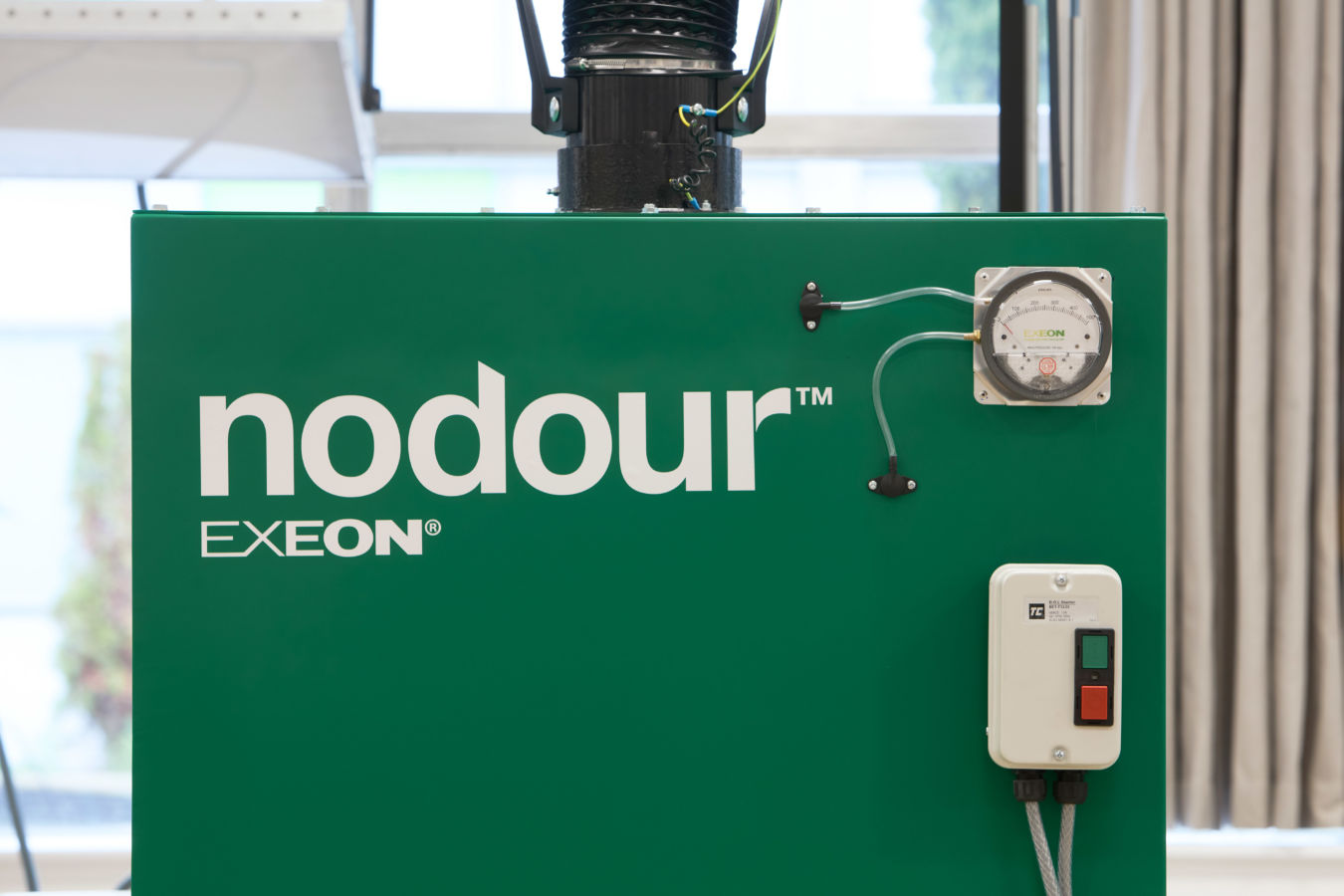 Green machine with the text "nodour™ EXEON®," featuring a gauge and small control box with power buttons, situated in an indoor environment with blurred windows and curtains in the background.