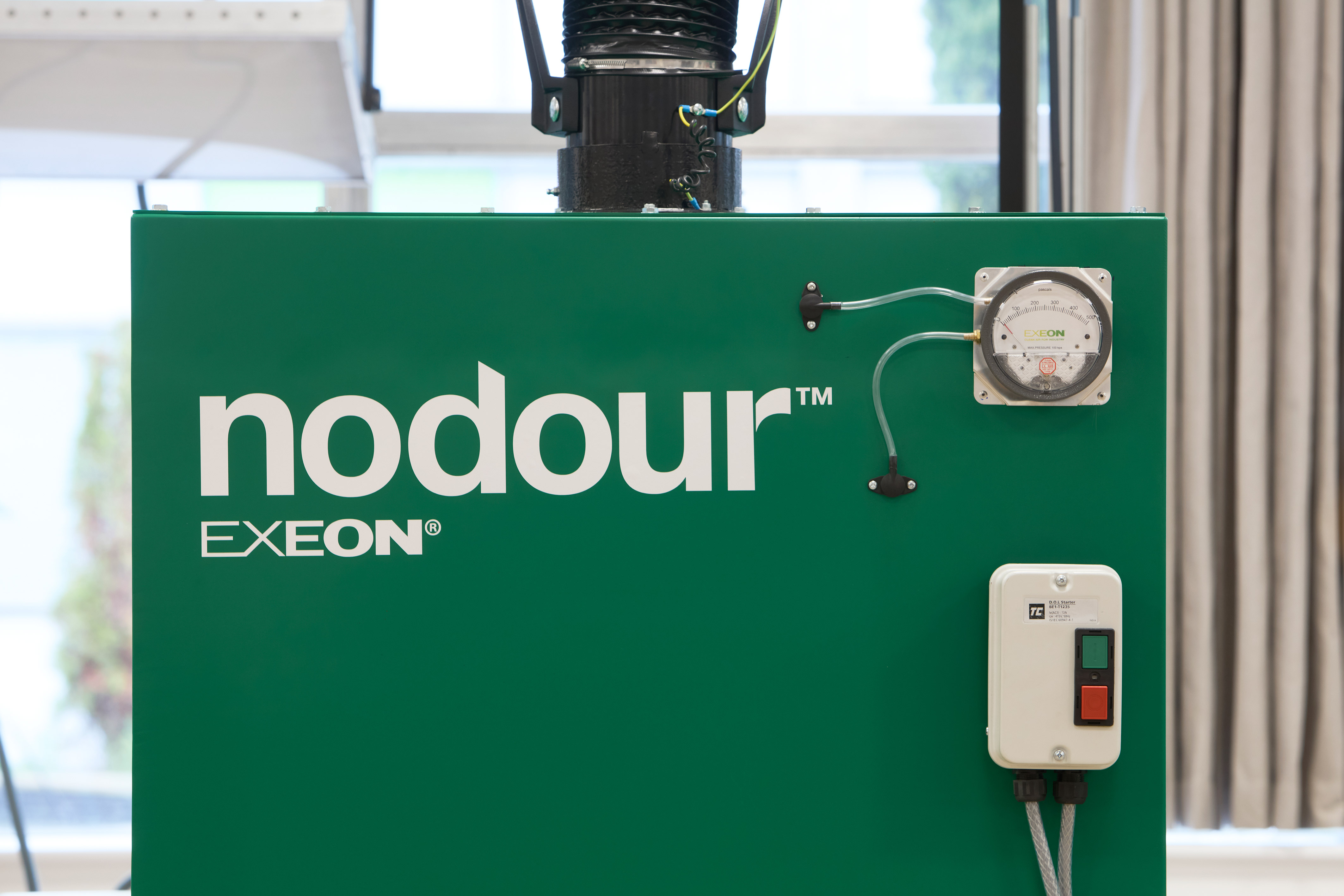 Green machine with the text "nodour™ EXEON®," featuring a gauge and small control box with power buttons, situated in an indoor environment with blurred windows and curtains in the background.