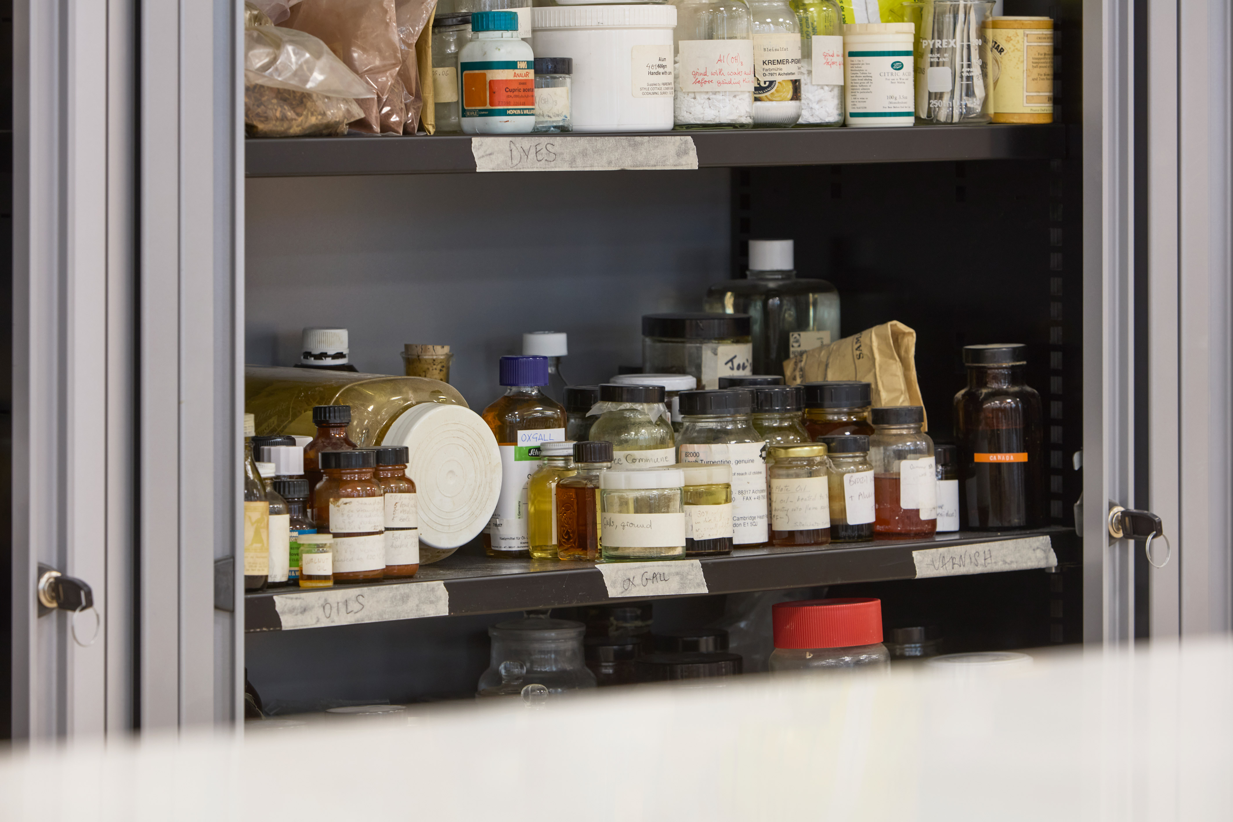 Shelves filled with various labeled jars and bottles of chemicals, including "Dyes," "Oils," and "Varnish," stored in a cabinet within a laboratory environment.