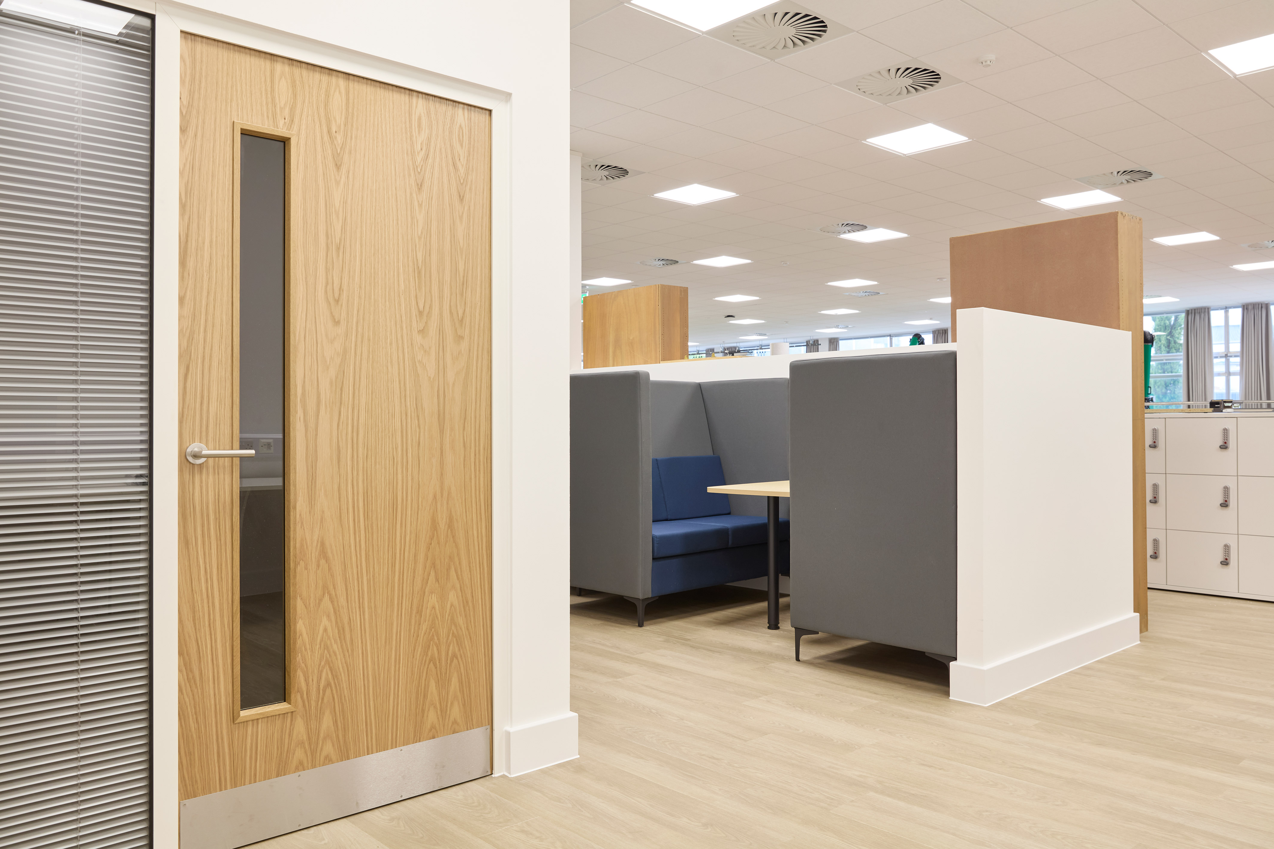 Wooden door stands closed with a long vertical window, adjacent to an office space with blue-gray booth seating and lockers, under a white ceiling with recessed lighting.