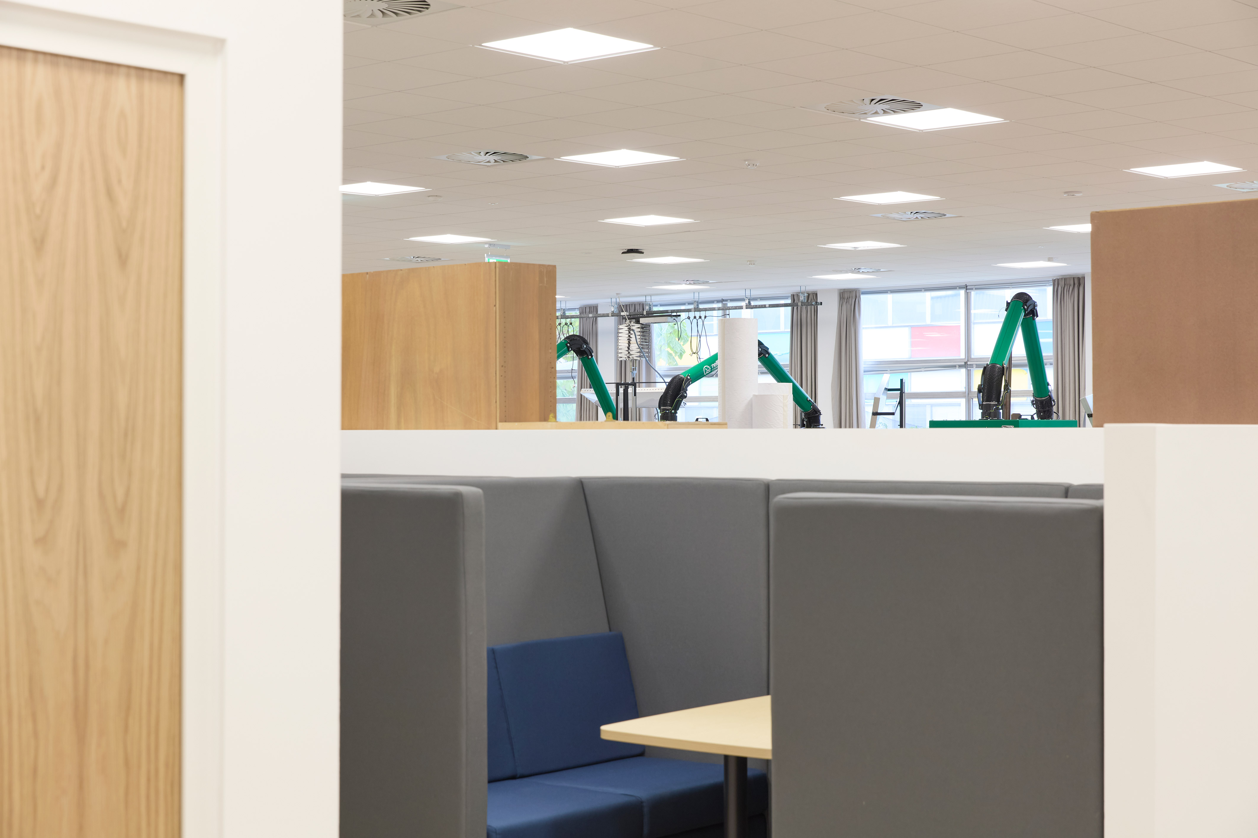 A gray meeting booth with a wooden table in an office space; large windows and visible green robotic arms suggest a modern, open-plan environment.