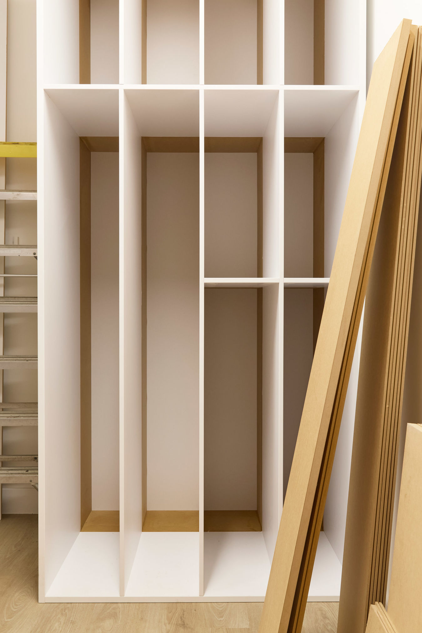 Partially assembled shelving unit in a room under construction, with wood panels leaning against the wall and a yellow step ladder in the background.