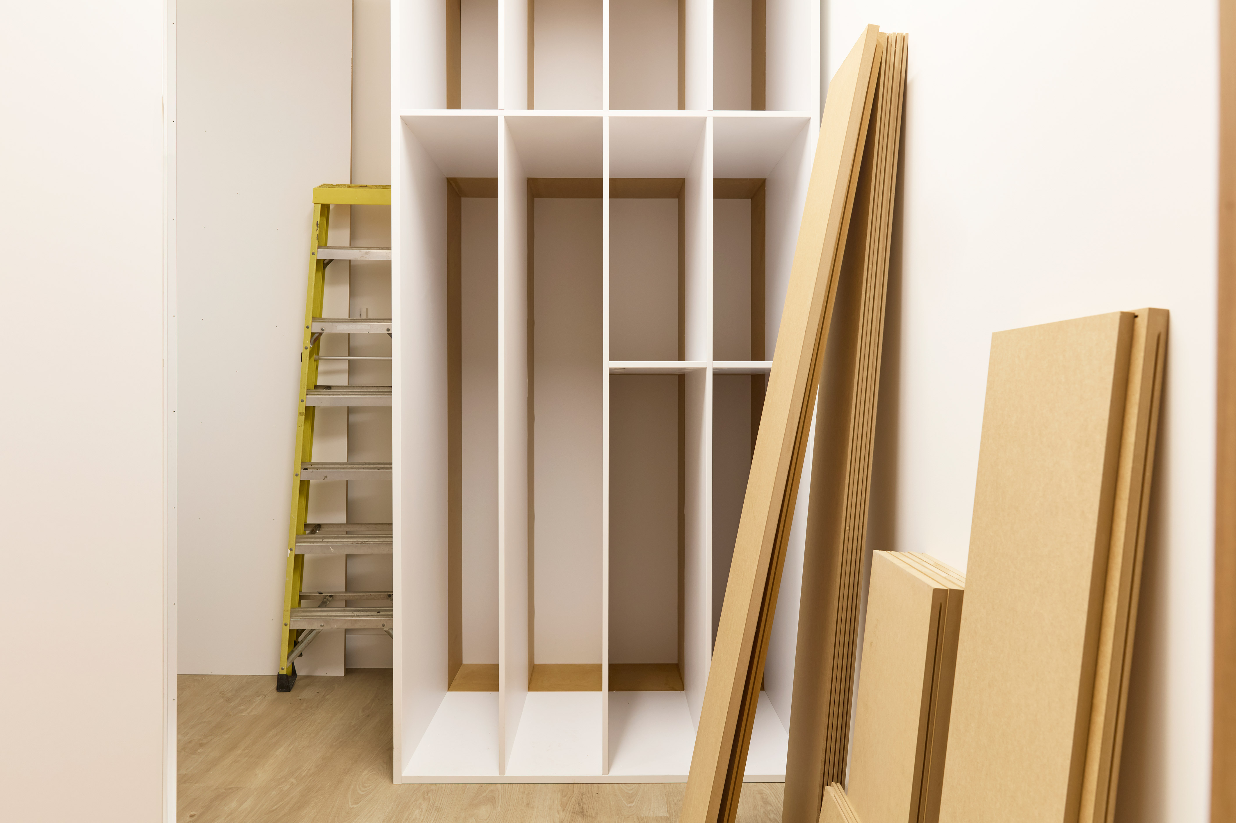 Partially assembled shelving unit in a room under construction, with wood panels leaning against the wall and a yellow step ladder in the background.