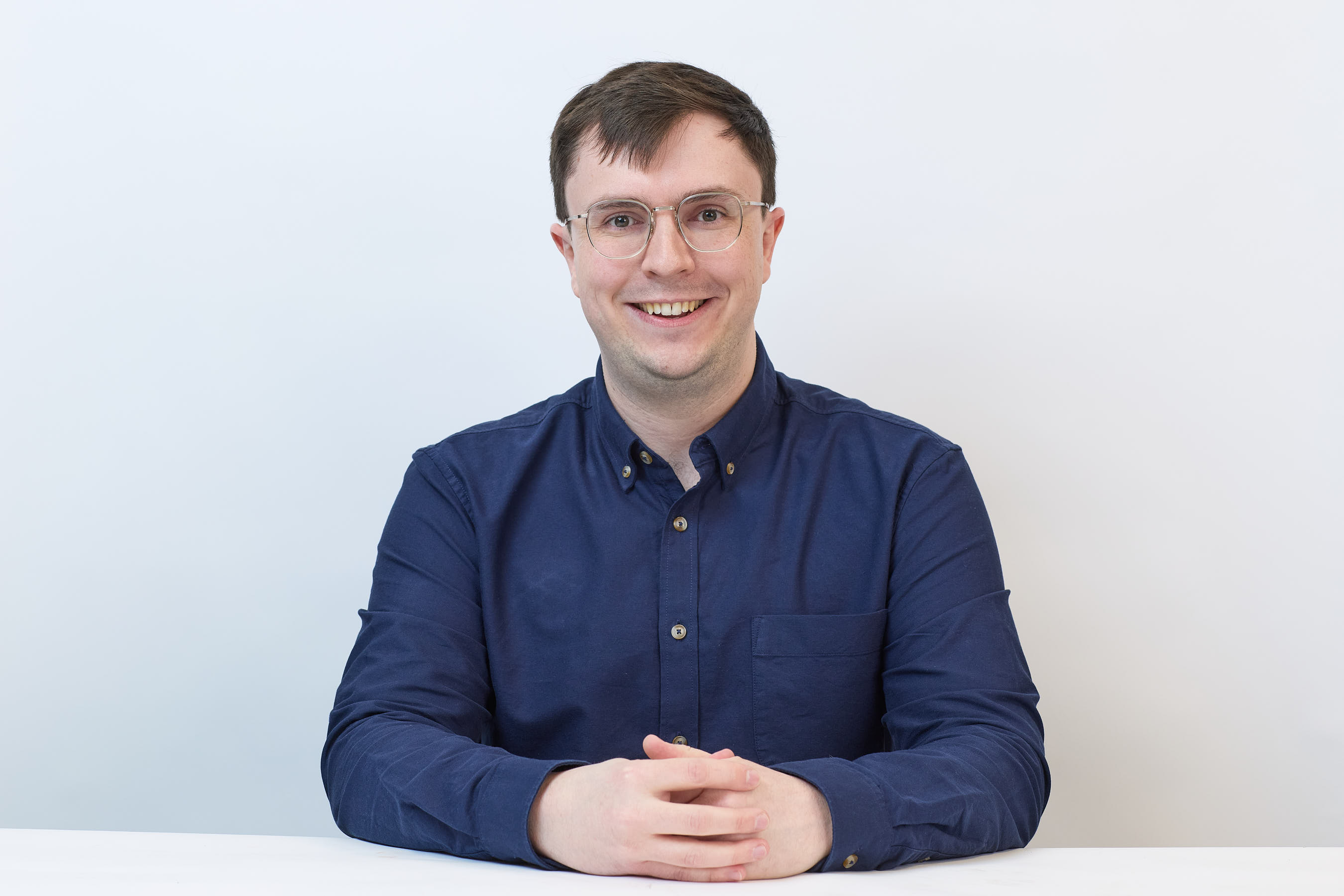 A smiling person wearing glasses and a blue button-up shirt sits with hands clasped on a white table against a plain white background.