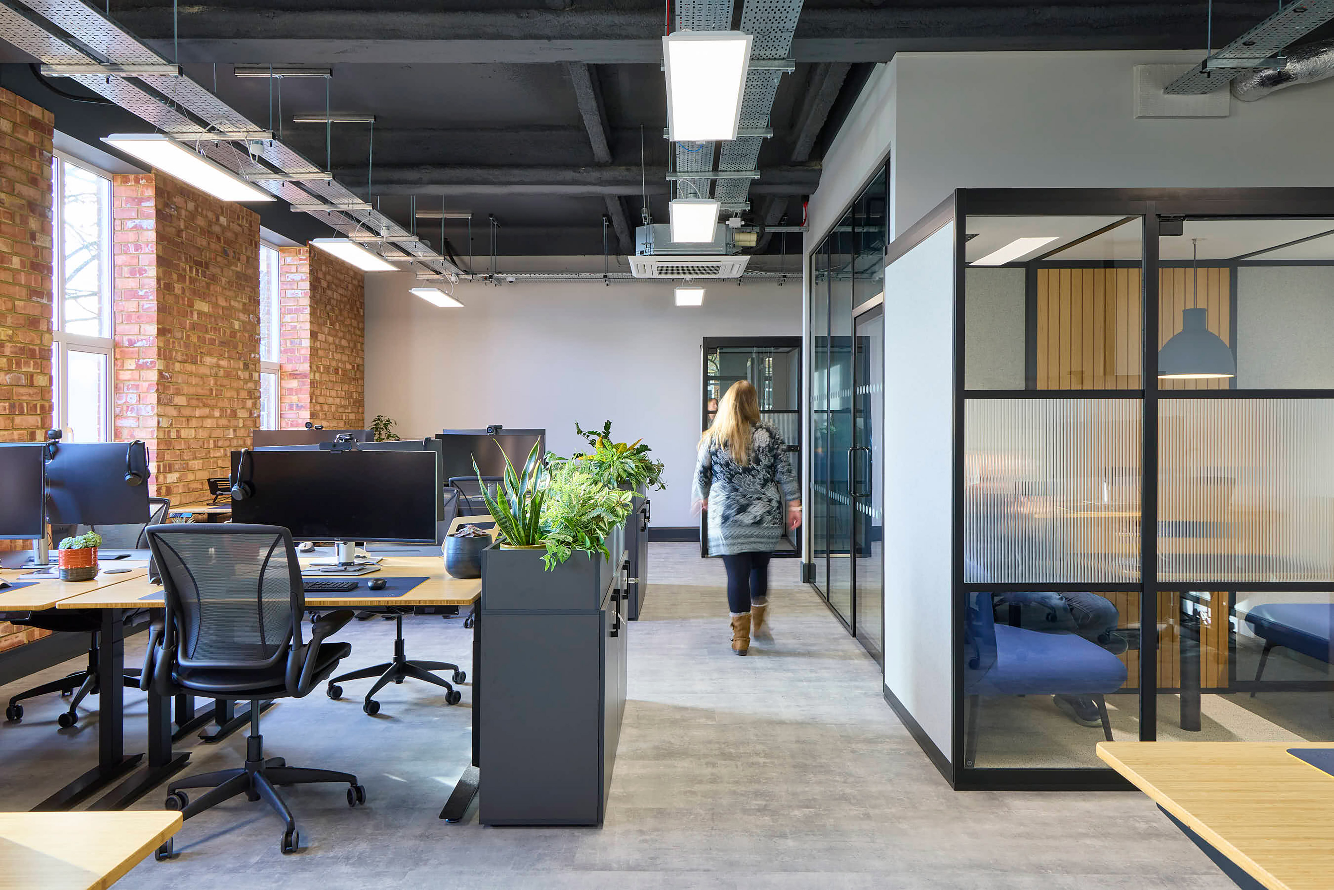 Open-plan office with workstations and computers; a person walks through, surrounded by brick walls, glass partitions, and overhead lighting; potted plants adorn desk areas.