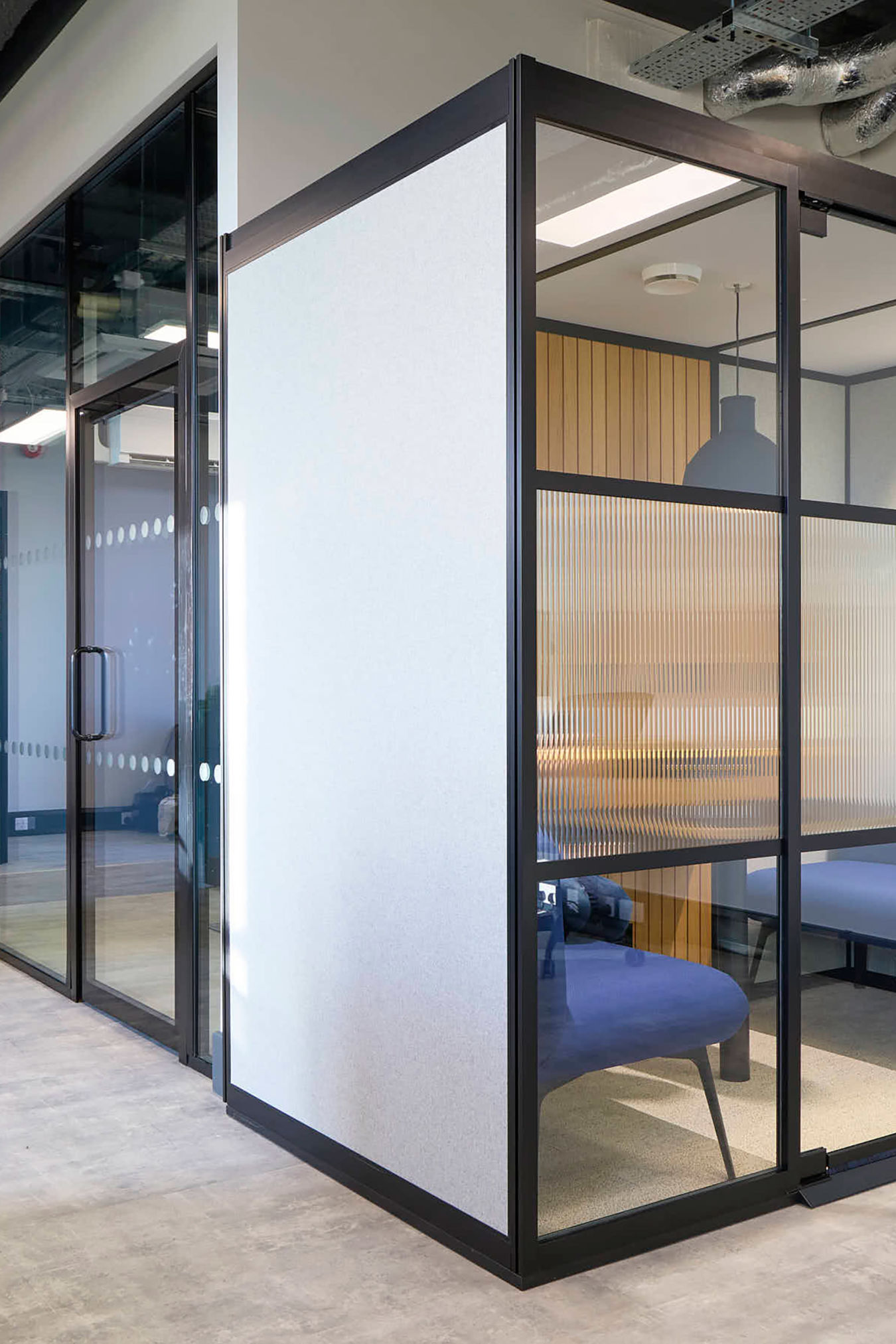 Modern office partitions with glass walls and black frames, enclosing blue seating areas; features exposed ceilings, overhead lighting, and a planter with green foliage in the foreground.
