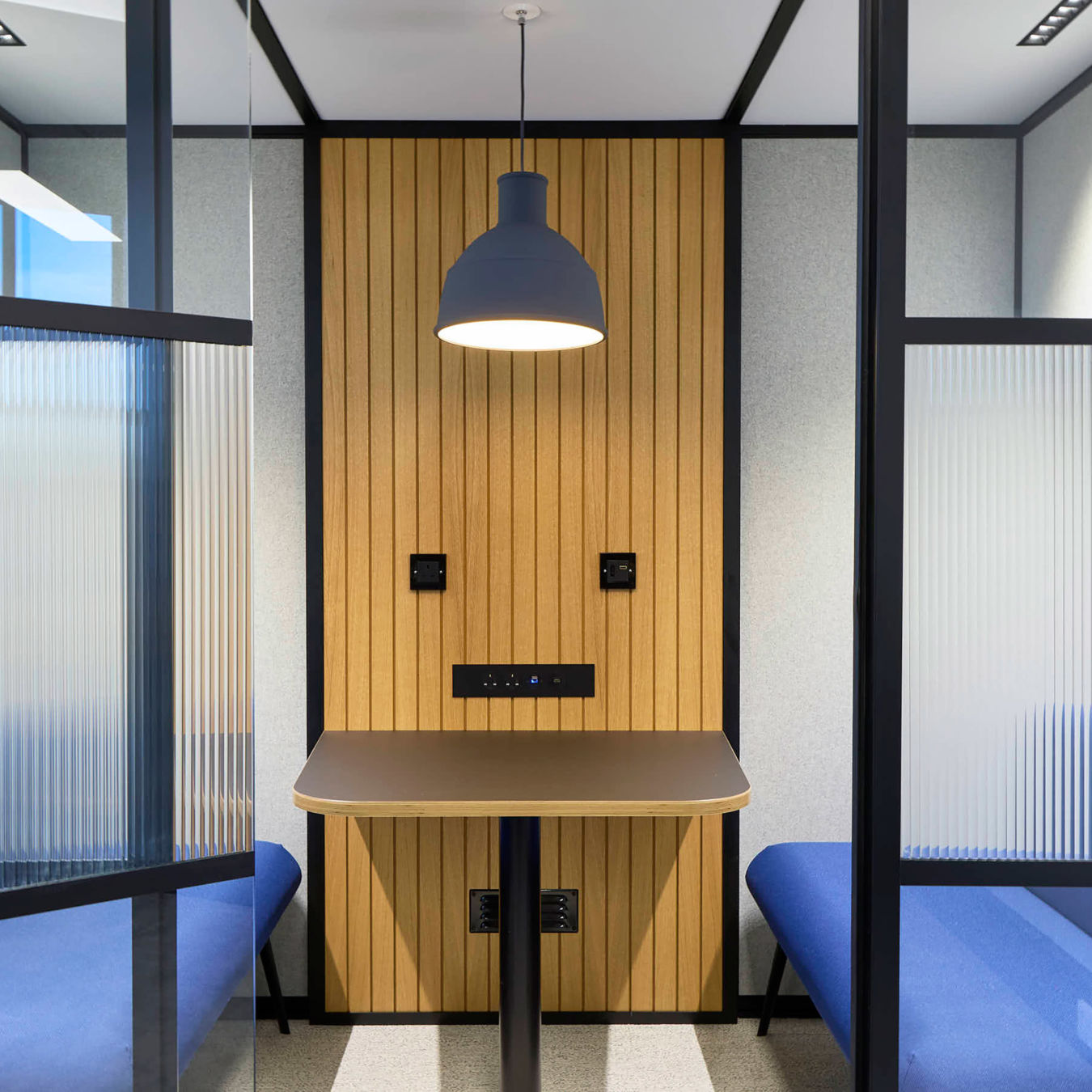 Modern office booth with blue seating and wooden paneling, separated by frosted glass. A black pendant light hangs over a central table, creating a cozy, focused atmosphere