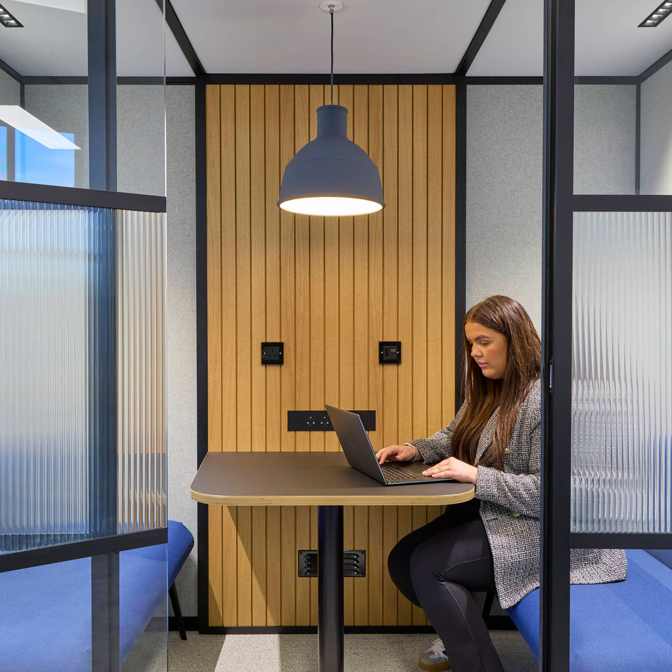 A woman works on a laptop at a table in a modern office booth, featuring wooden paneling, frosted glass dividers, blue benches, and overhead lighting.