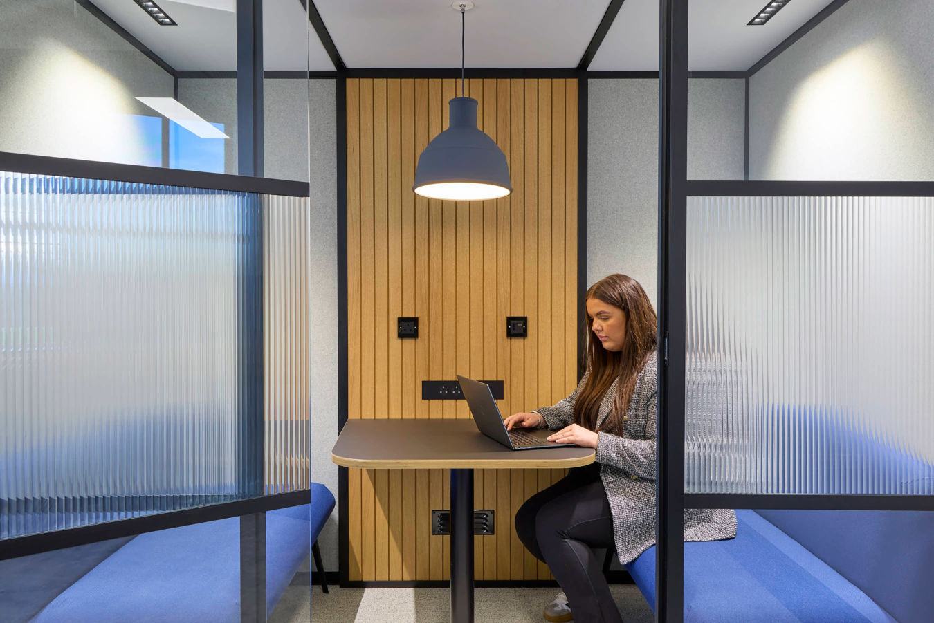 A woman works on a laptop at a table in a modern office booth, featuring wooden paneling, frosted glass dividers, blue benches, and overhead lighting.