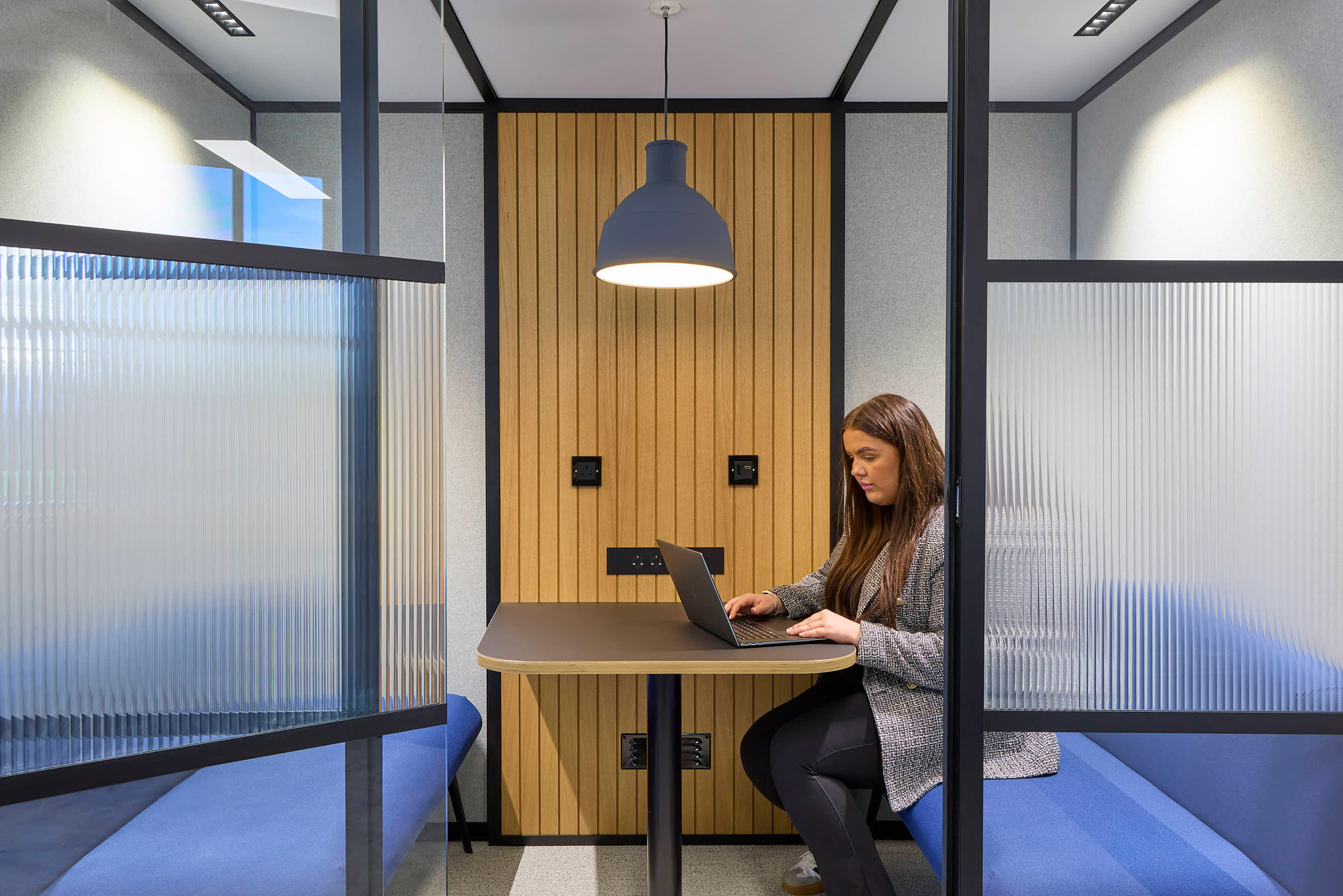 A woman works on a laptop at a table in a modern office booth, featuring wooden paneling, frosted glass dividers, blue benches, and overhead lighting.