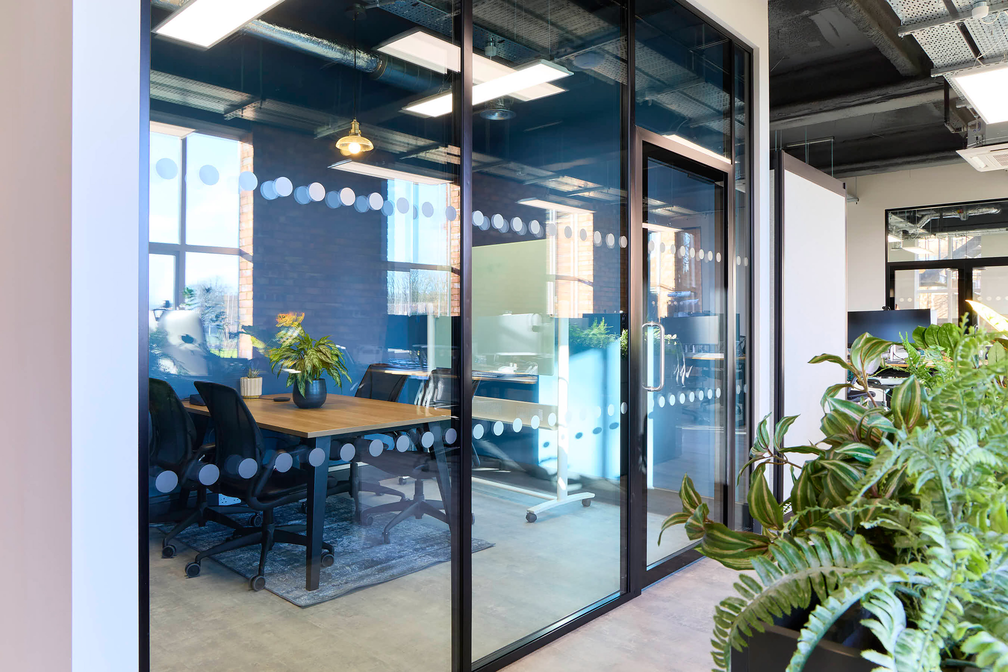 Glass-walled office room contains a wooden table surrounded by black chairs, with potted plants and overhead lighting, situated in a modern open office environment featuring natural light and greenery.