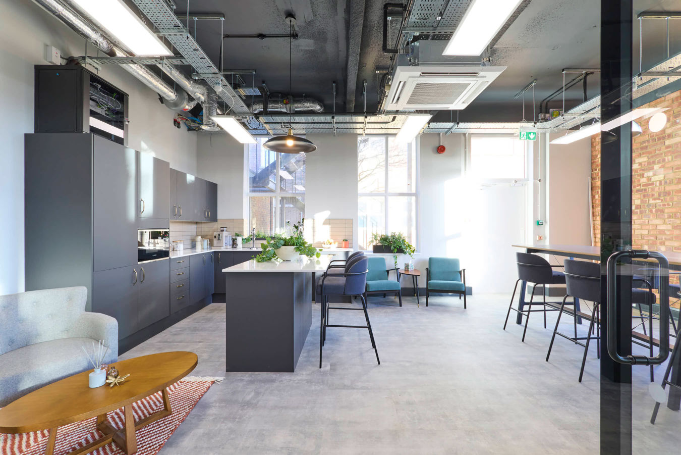 Modern office kitchen with sleek dark cabinets, a central island, and tall stools. Large windows let in natural light, creating a bright, airy feel.