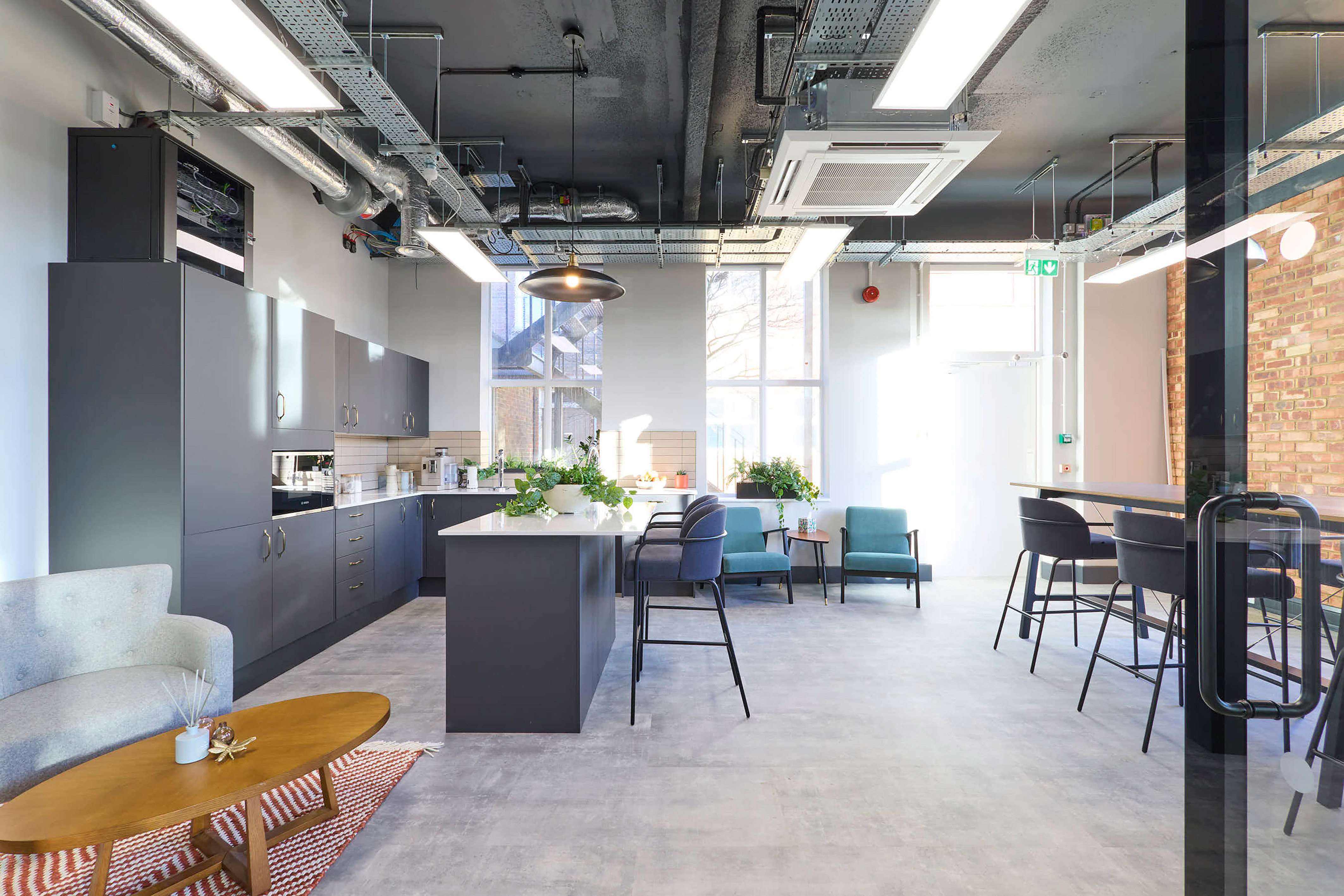 Modern office kitchen with sleek dark cabinets, a central island, and tall stools. Large windows let in natural light, creating a bright, airy feel.