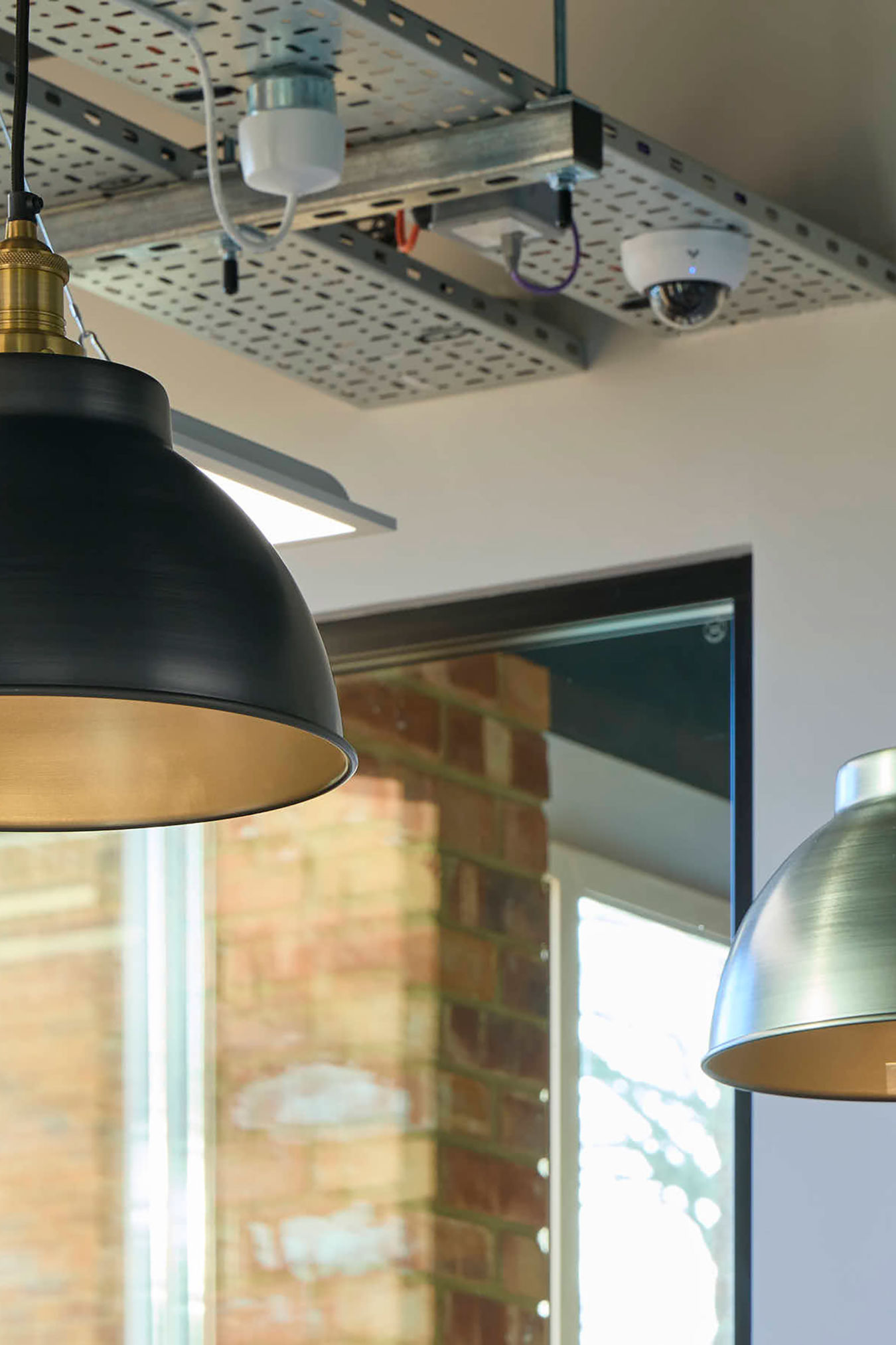 Hanging light fixtures illuminate an indoor space with exposed ductwork and a security camera on the ceiling; a window reveals a brick wall outside.