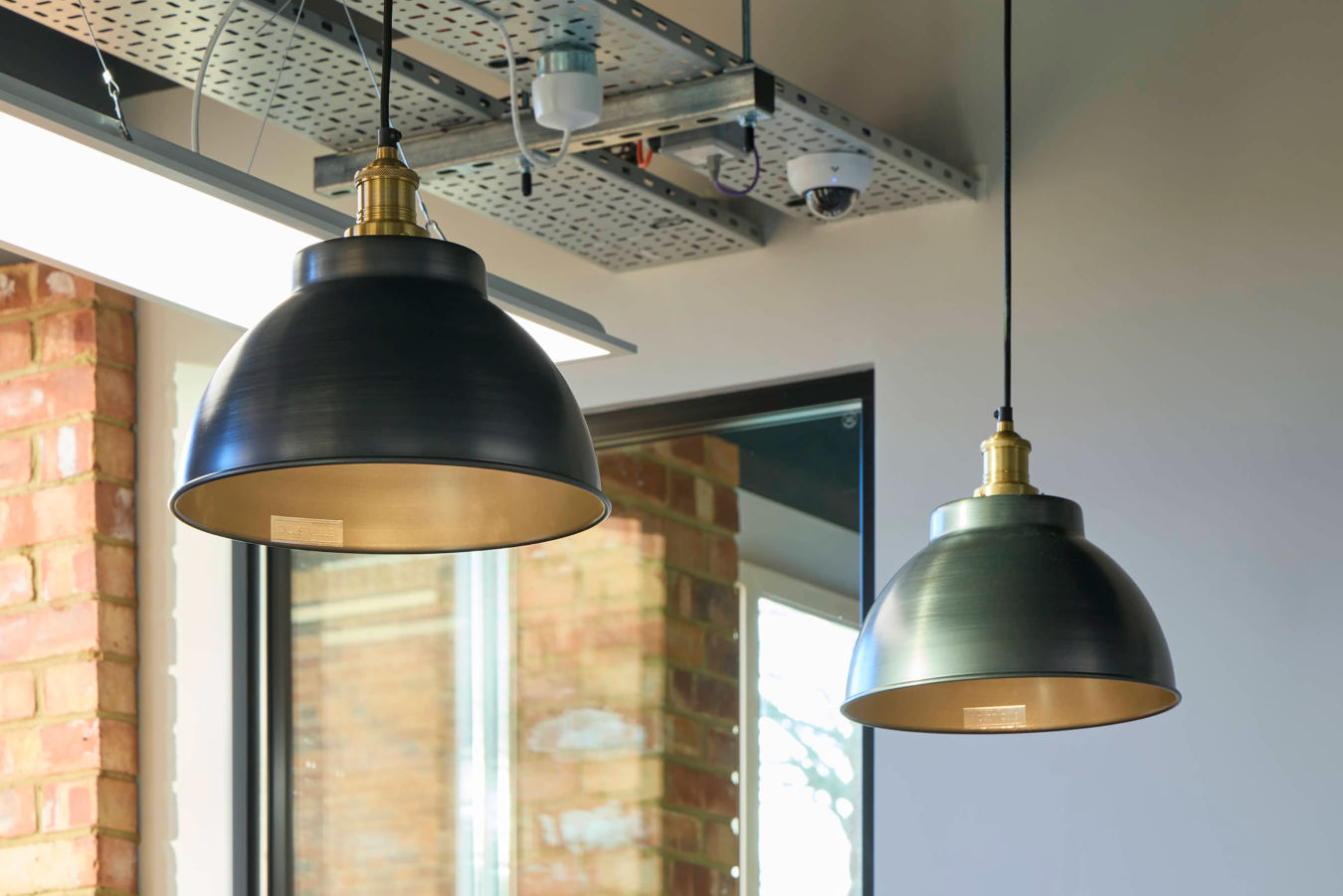 Hanging light fixtures illuminate an indoor space with exposed ductwork and a security camera on the ceiling; a window reveals a brick wall outside.