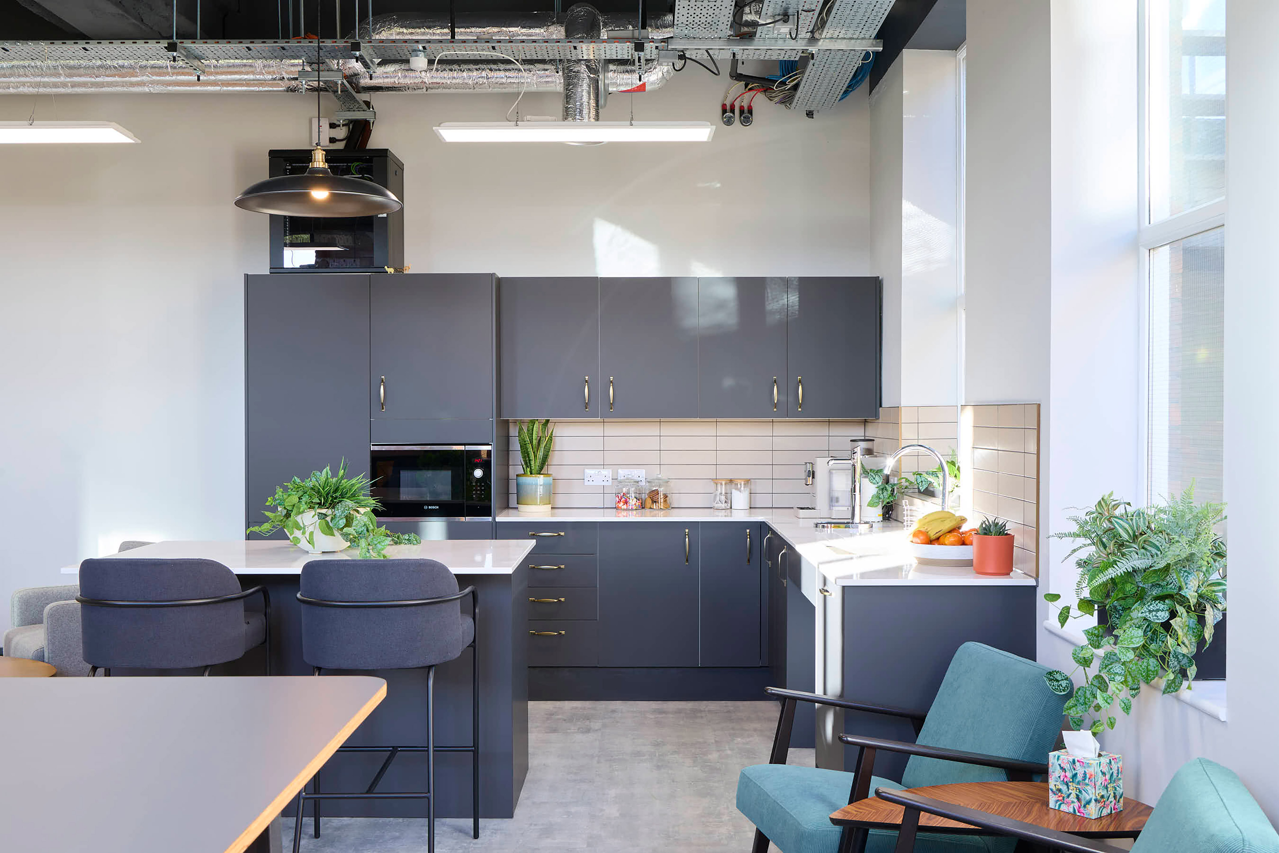 Modern kitchen area featuring dark cabinets, a wall-mounted oven, and a white countertop adorned with plants and fruit. Surrounding context includes grey bar stools, teal chairs, and large windows.
