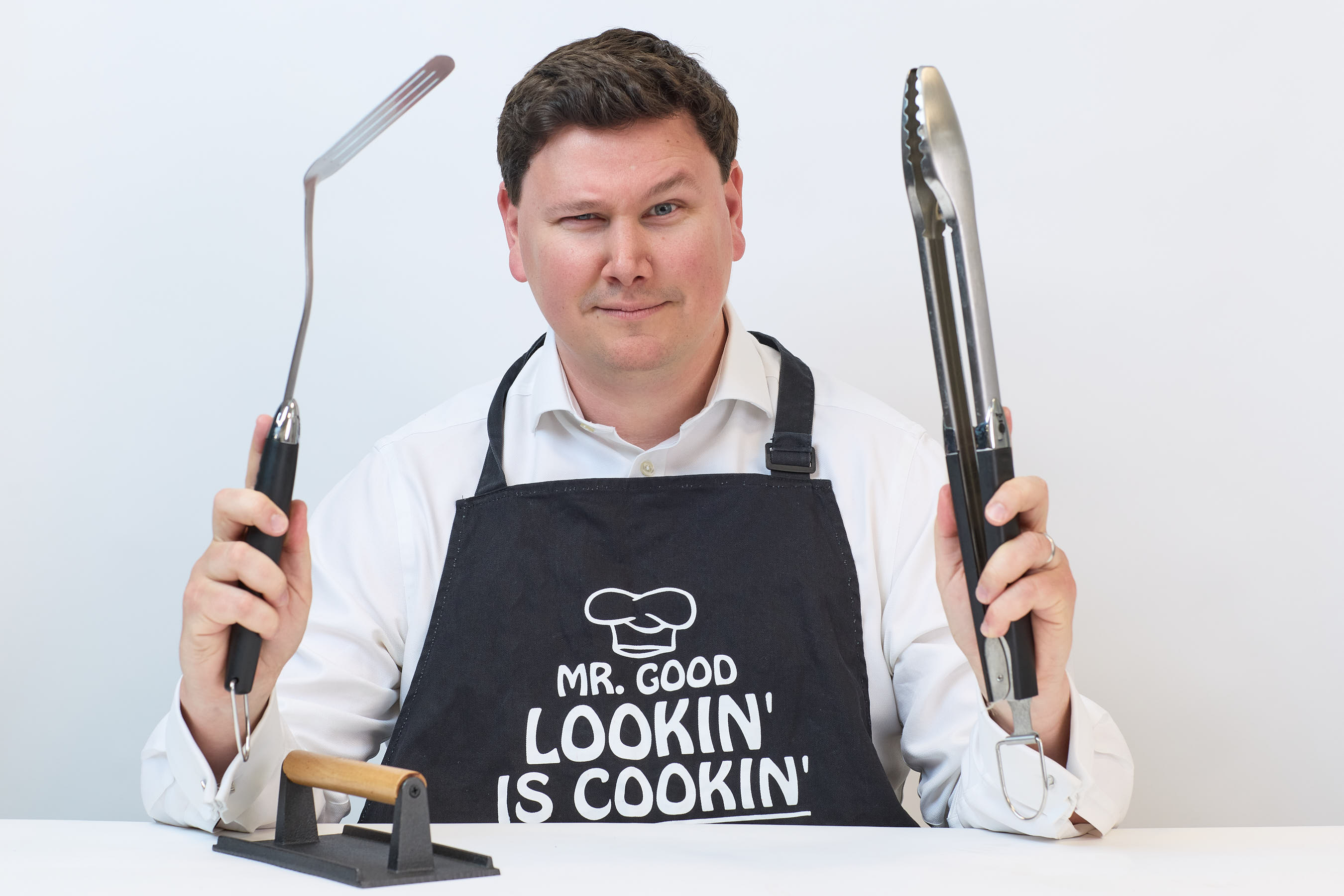 A person wearing an apron reading "MR. GOOD LOOKIN' IS COOKIN'" holds grilling tools, smiling, at a table, against a plain white background.
