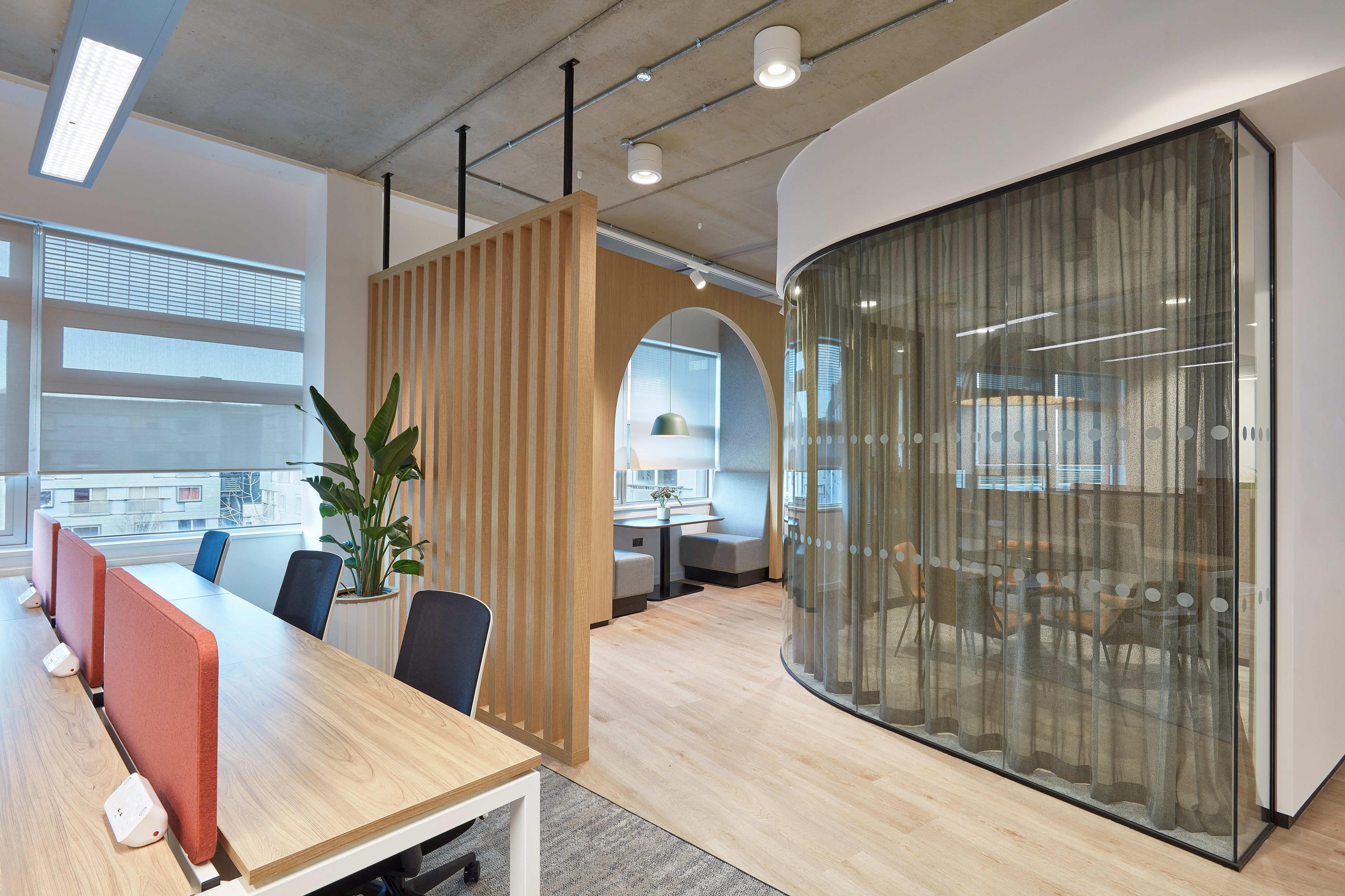 Modern office space featuring a long wooden desk with orange dividers and chairs. Curved glass meeting room with sheer curtains, next to a wooden slatted partition. Bright, natural lighting.