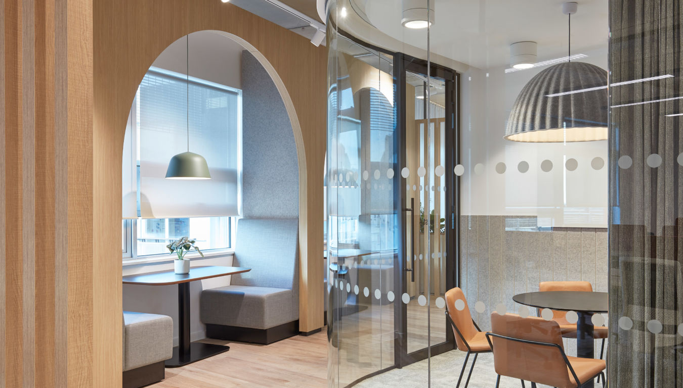 office with a cozy seating nook under an arched wooden frame, featuring gray cushions, a hanging lamp, and a small plant. Curved glass wall encloses a meeting space, adding a sleek, inviting atmosphere