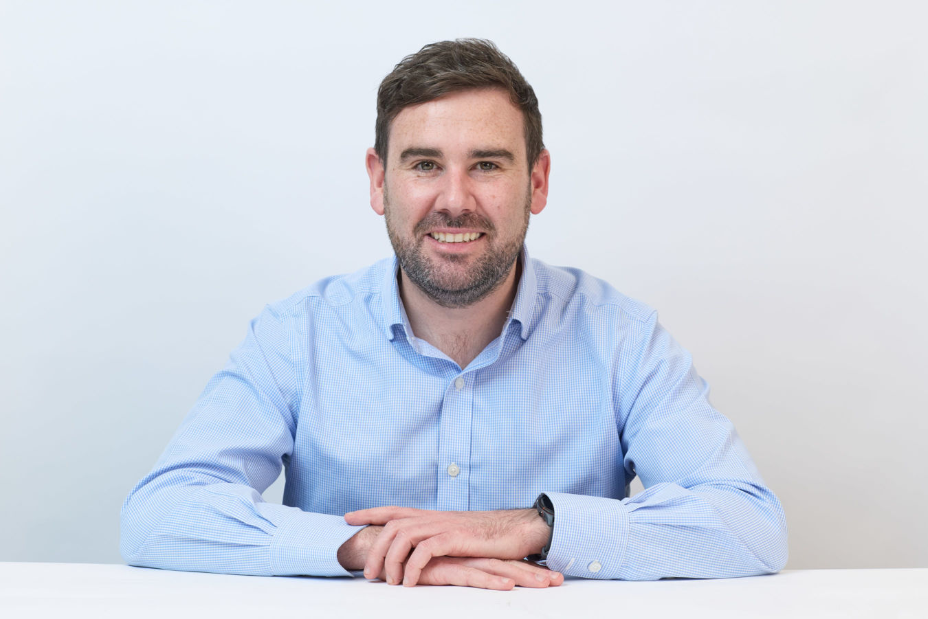 Man in a light blue checkered shirt, seated with arms crossed and smiling, against a plain white background.