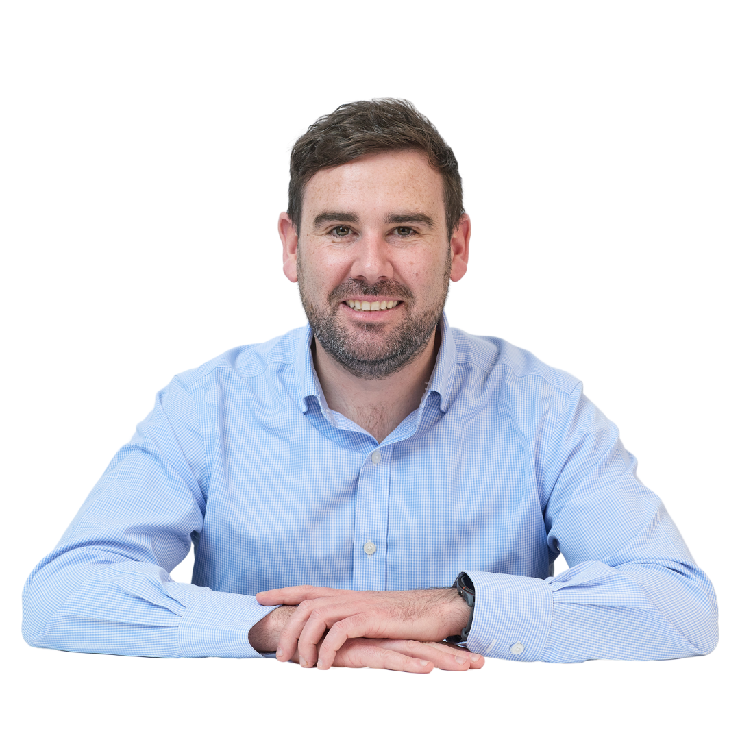 A man in a light blue shirt smiling and resting his arms on a surface against a plain white background.