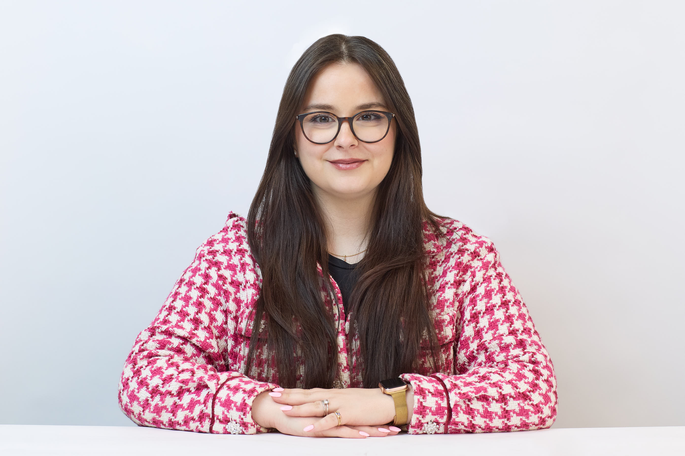 A person with long hair and glasses is sitting, resting hands on a surface, wearing a red patterned jacket. The background is plain white, emphasizing the individual.