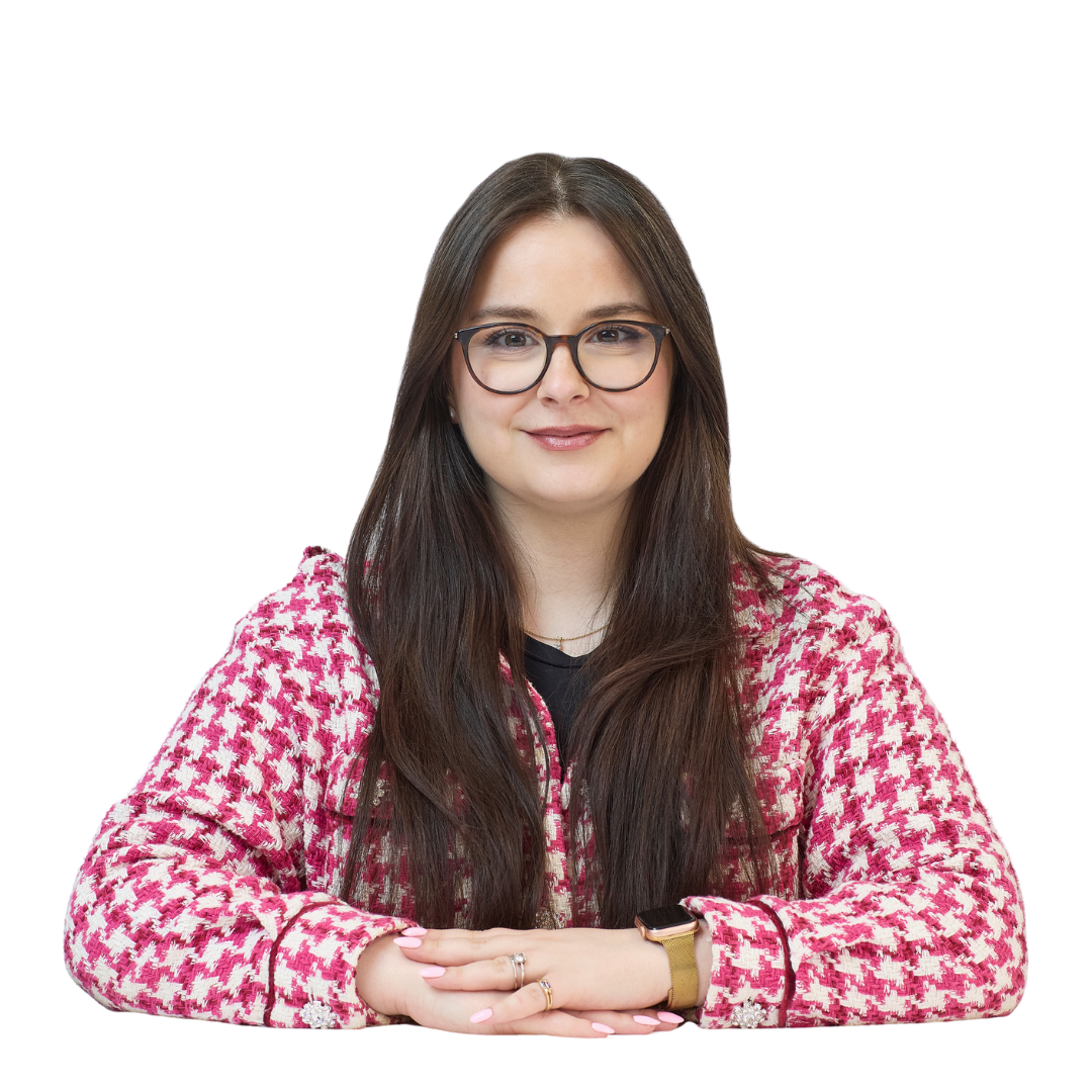 A person with long hair and glasses sits with folded hands on a white surface, wearing a pink and white checkered jacket, against a plain light background.