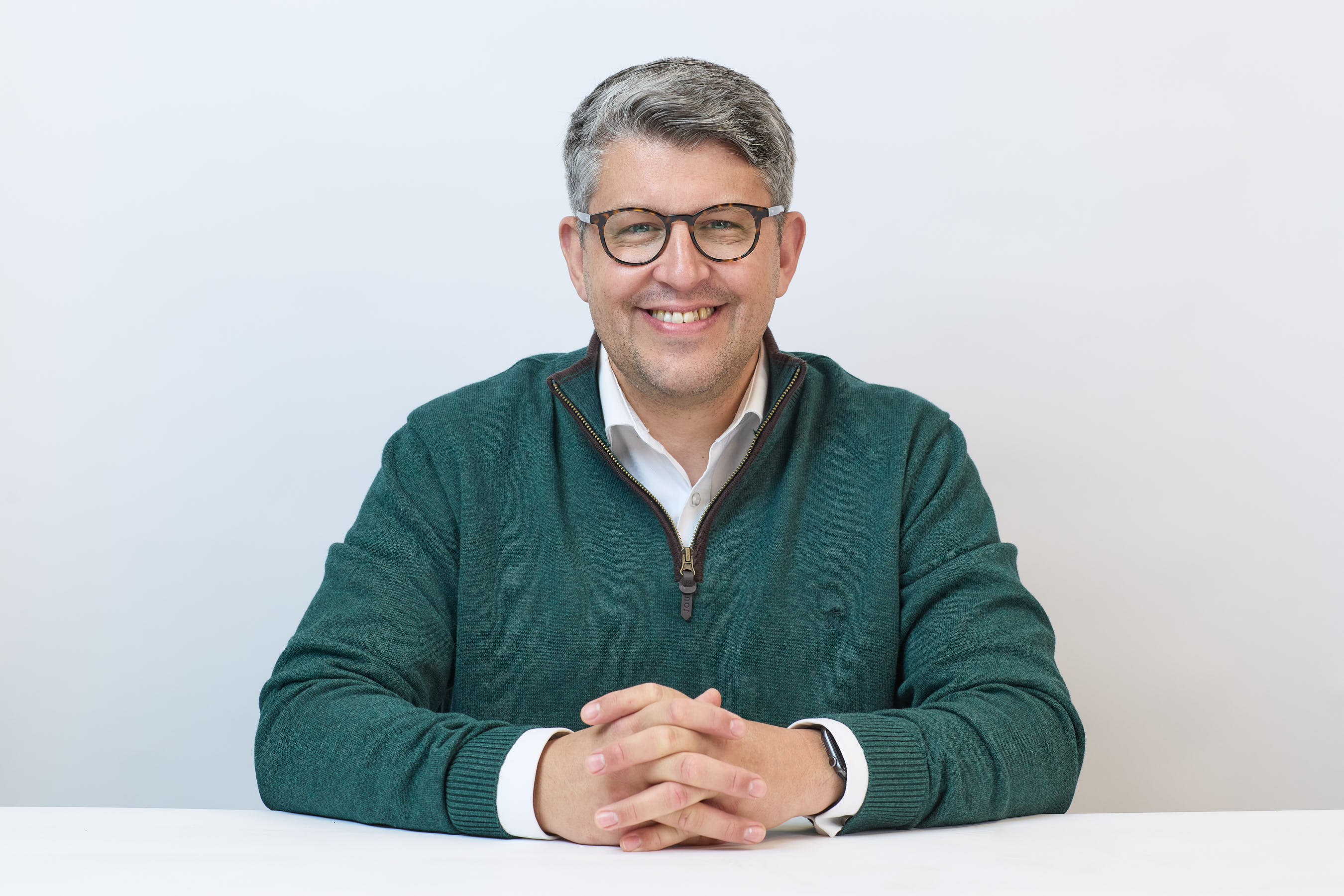 A person wearing glasses and a green pullover smiles while sitting at a white table against a plain background.