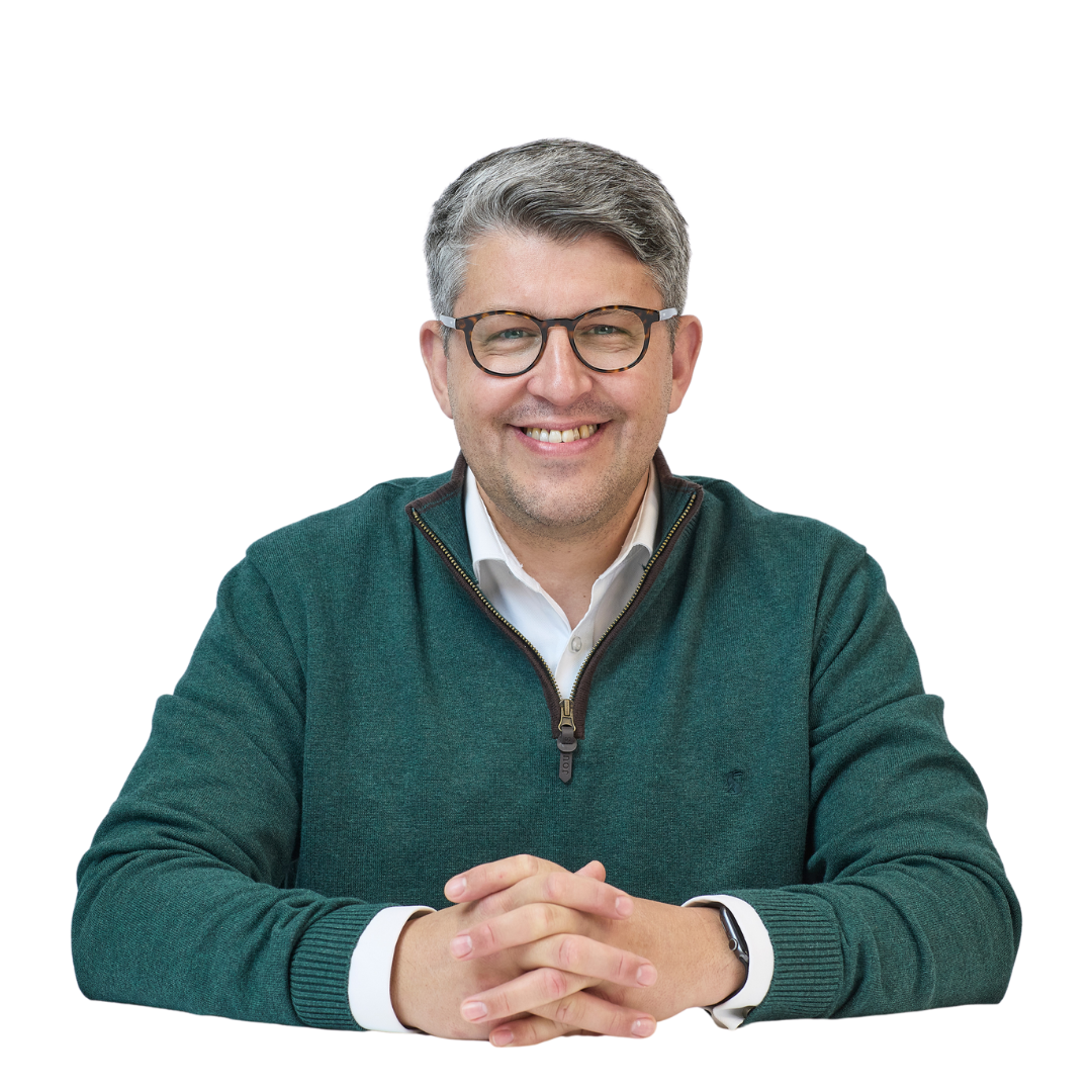 A person wearing a green sweater and glasses smiles while seated with hands clasped, set against a plain white background.