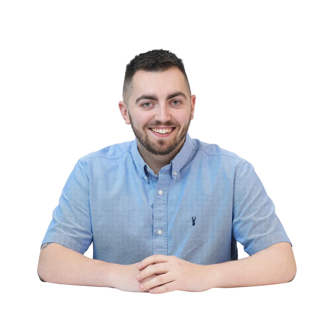 A man with short hair and a beard smiles while sitting at a table, wearing a light blue button-up shirt, on a plain white background.