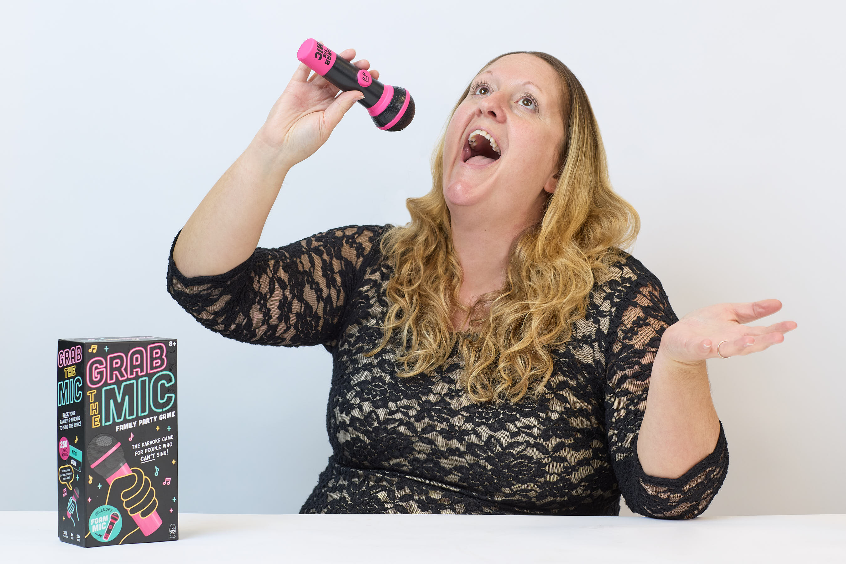A person holds a toy microphone as if singing, beside a box labeled "GRAB THE MIC: THE FAMILY PARTY GAME" on a table, against a plain background.