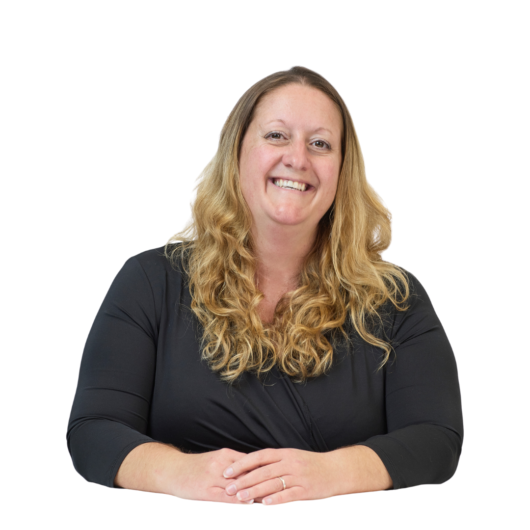 A smiling person with long, wavy hair sits at a table, wearing a black top, hands gently clasped, against a plain white background.