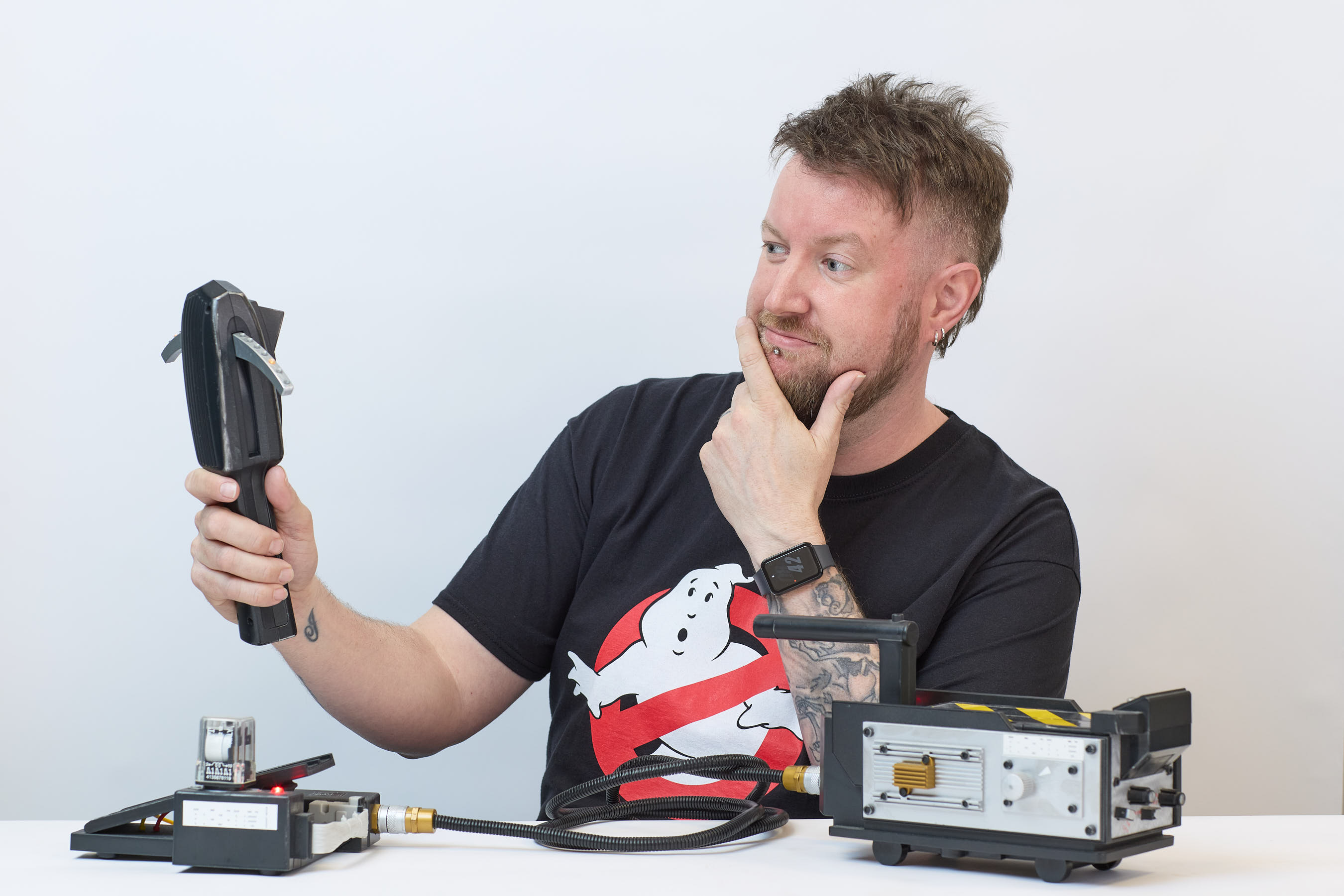 A person holds a black device inquisitively while seated at a table with electronic gadgets. They wear a Ghostbusters t-shirt and smartwatch, appearing thoughtful against a plain background.
