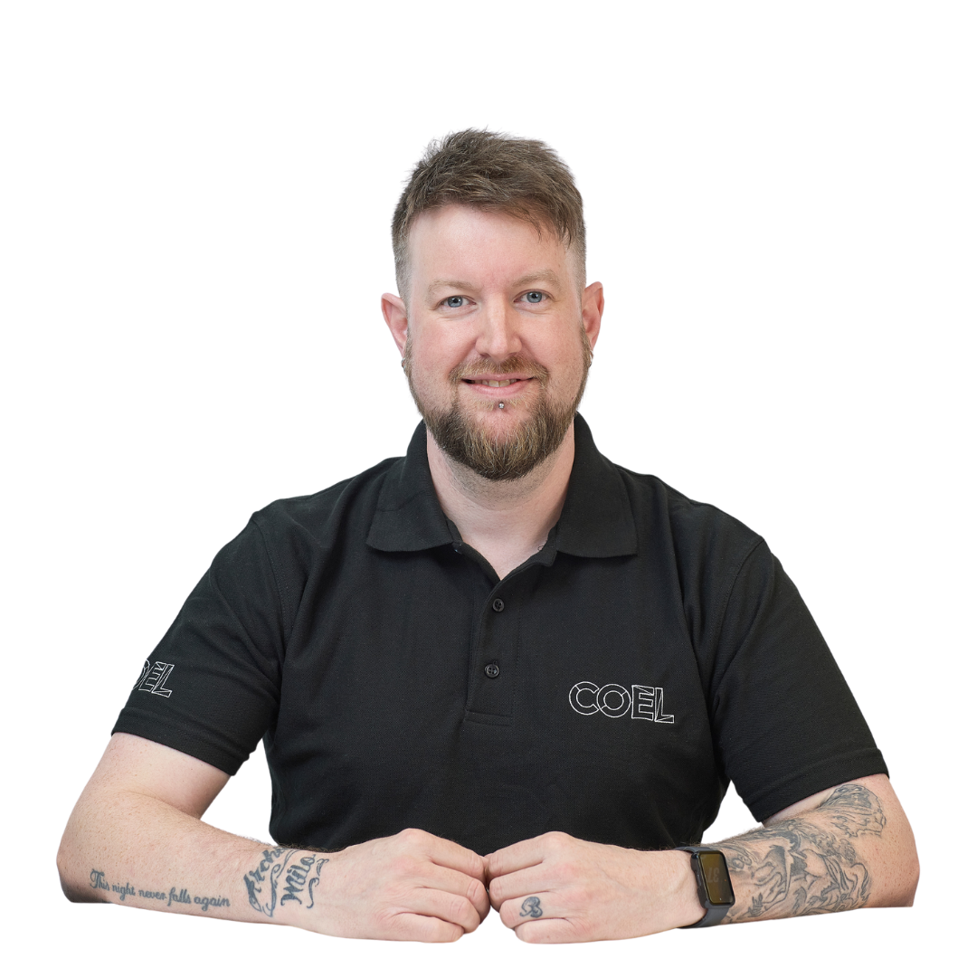 A person sits, smiling and wearing a black polo shirt with "COEL" on the chest and sleeve, displaying tattoos on their forearms, against a plain white background.