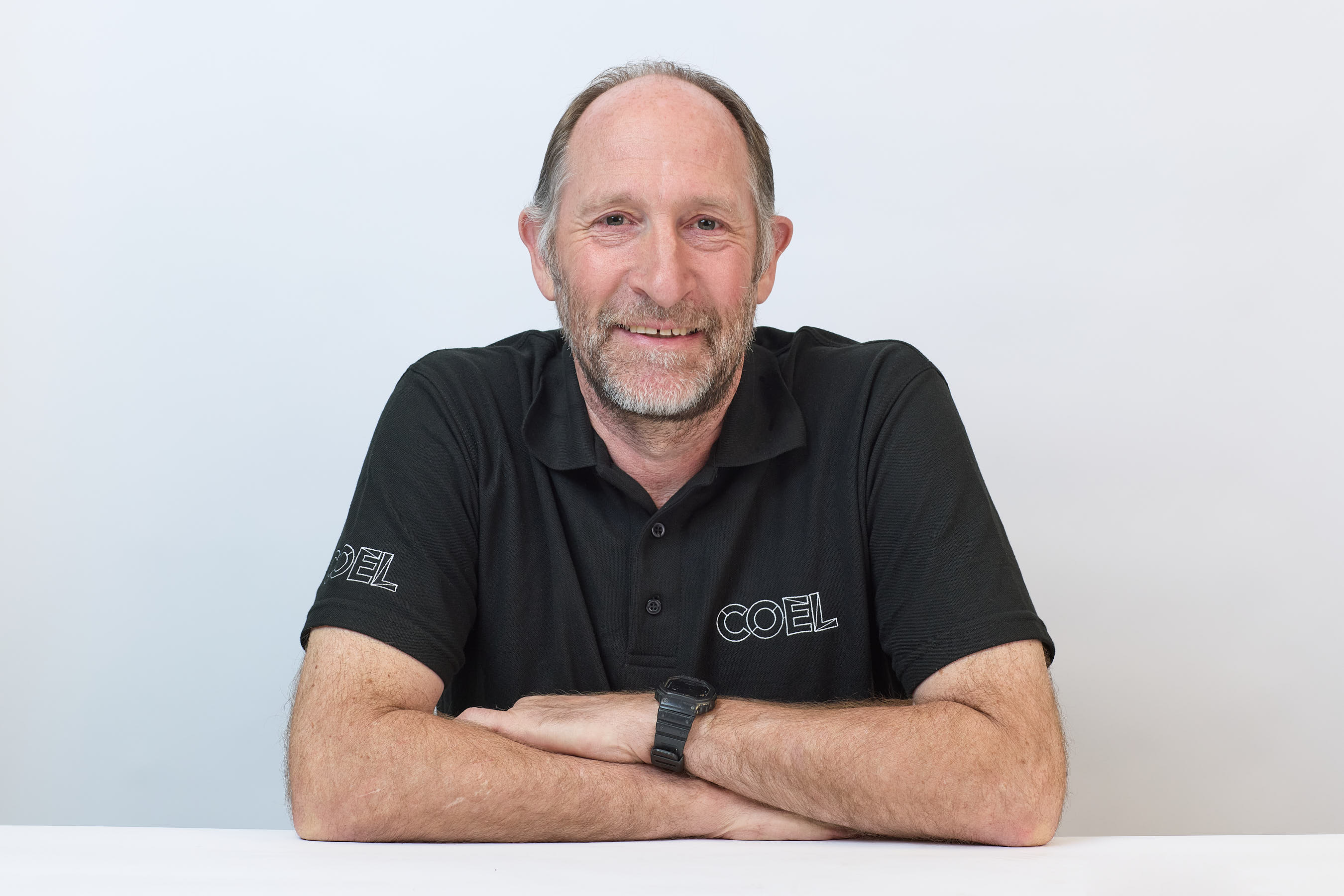 Man smiling with arms crossed, wearing a black polo shirt with "COEL" on the sleeve and chest, in front of a plain white background.