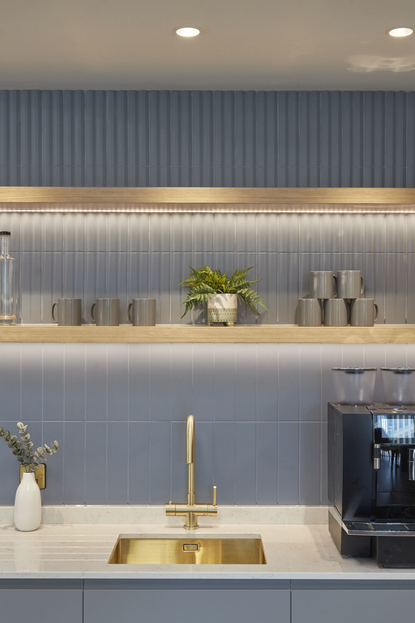 Modern kitchen area with a gold faucet over a rectangular sink, flanked by a coffee machine and a white vase with foliage. Shelves hold mugs and a potted plant, set against blue tiles.