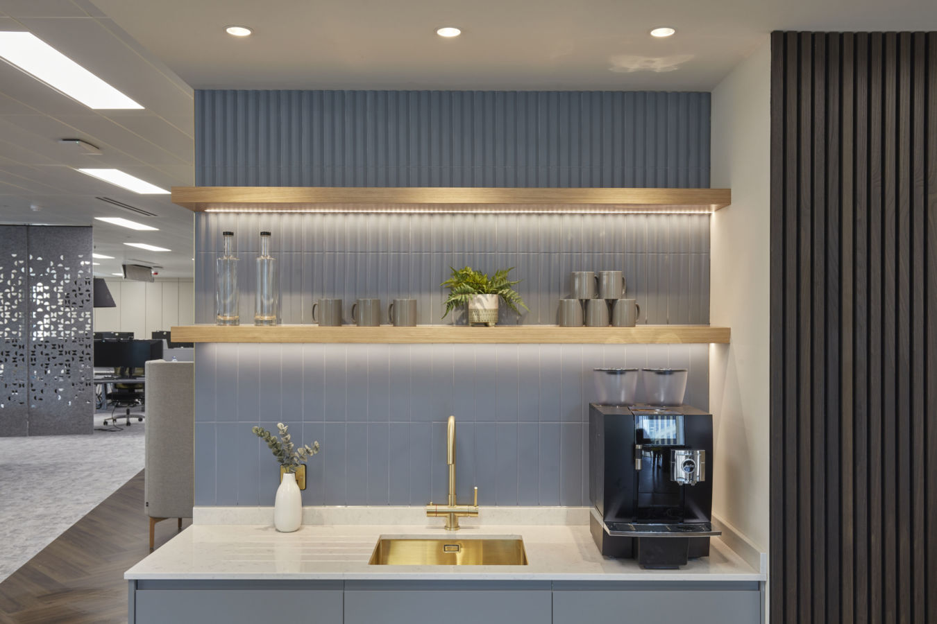 Modern kitchen area with a gold faucet over a rectangular sink, flanked by a coffee machine and a white vase with foliage. Shelves hold mugs and a potted plant, set against blue tiles.