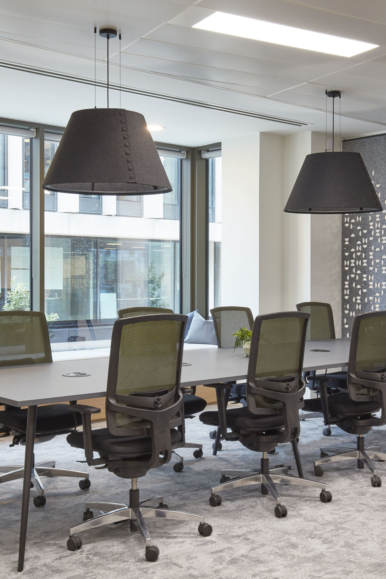 Modern conference room with large table, eight office chairs, and two pendant lights. Bright windows, lounge seating, and decorative wall panel. Minimalist style