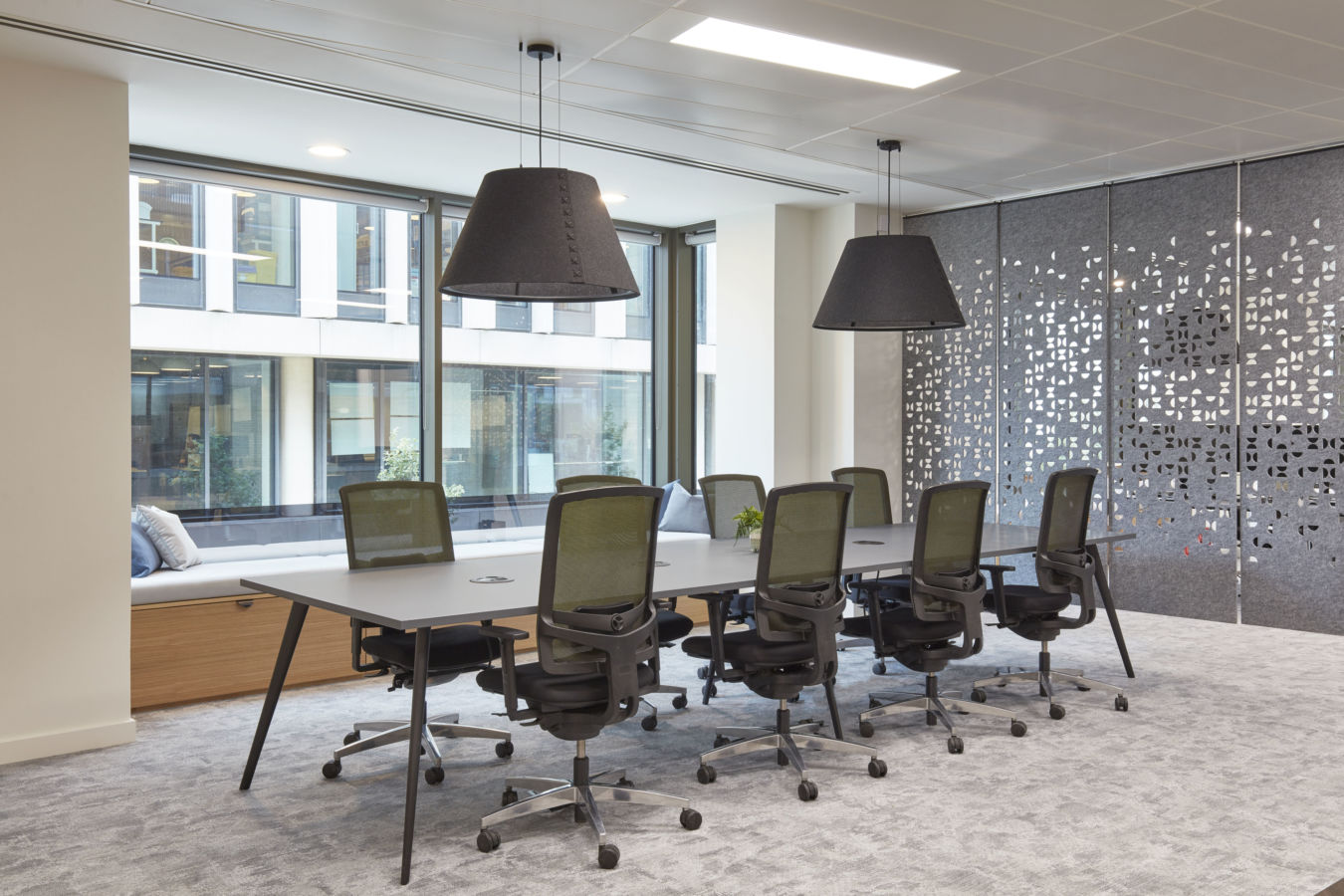 Modern conference room with large table, eight office chairs, and two pendant lights. Bright windows, lounge seating, and decorative wall panel. Minimalist style