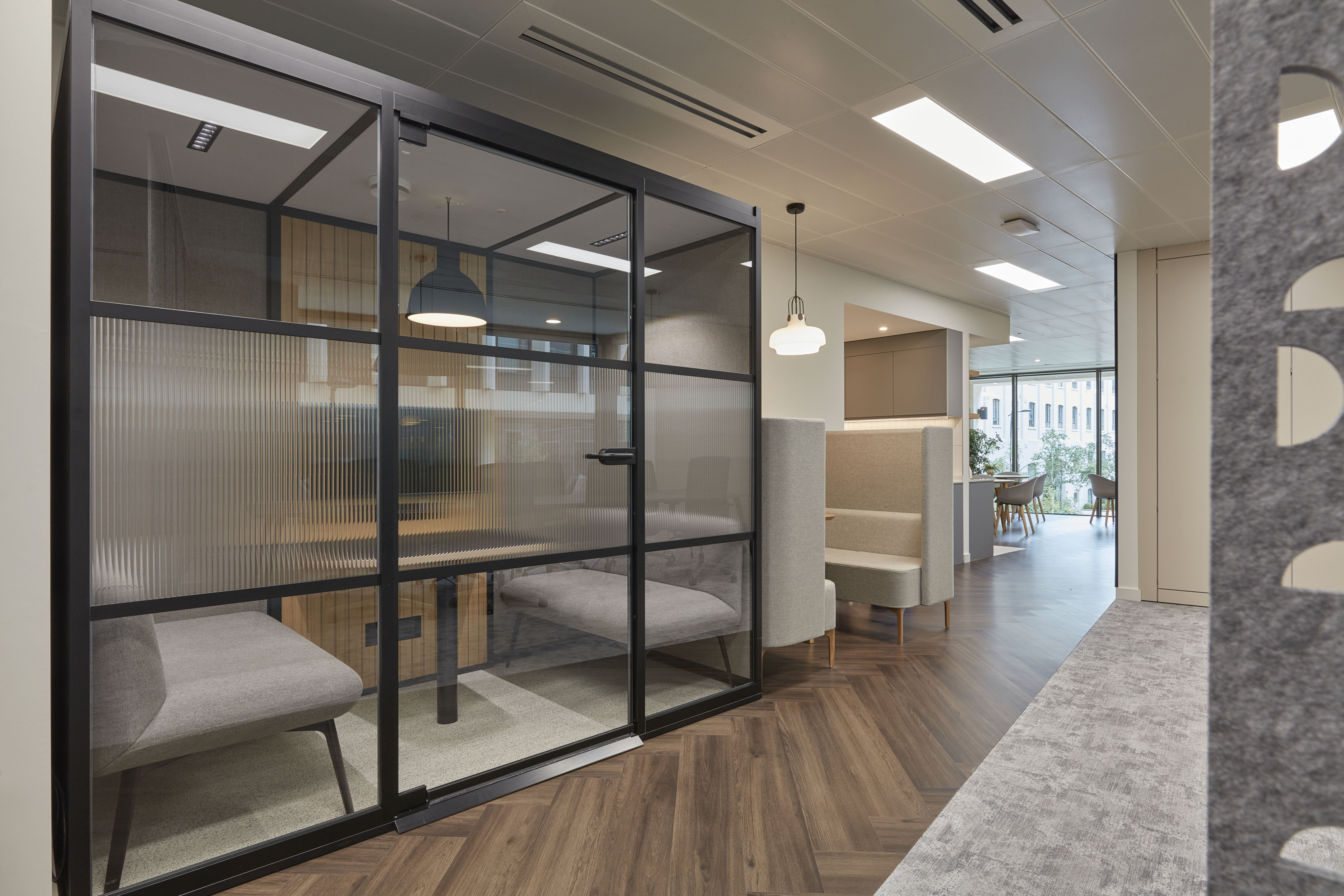 Office interior with glass-partitioned meeting booths, wood flooring, and soft seating, completed by COEL for Mishcon De Reya.