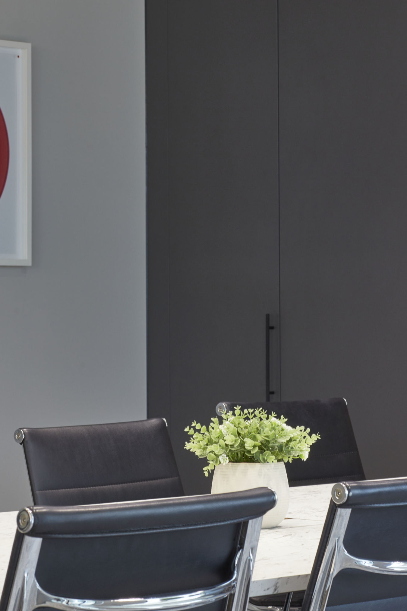 A potted plant sits on a white marble table surrounded by black leather chairs in a modern office setting with a partial view of a red artwork on the wall.