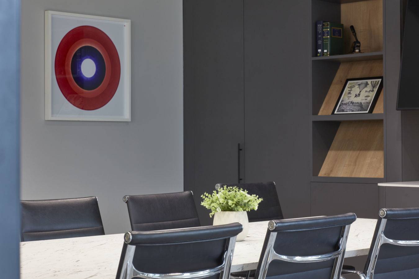 A potted plant sits on a white marble table surrounded by black leather chairs in a modern office setting with a partial view of a red artwork on the wall.