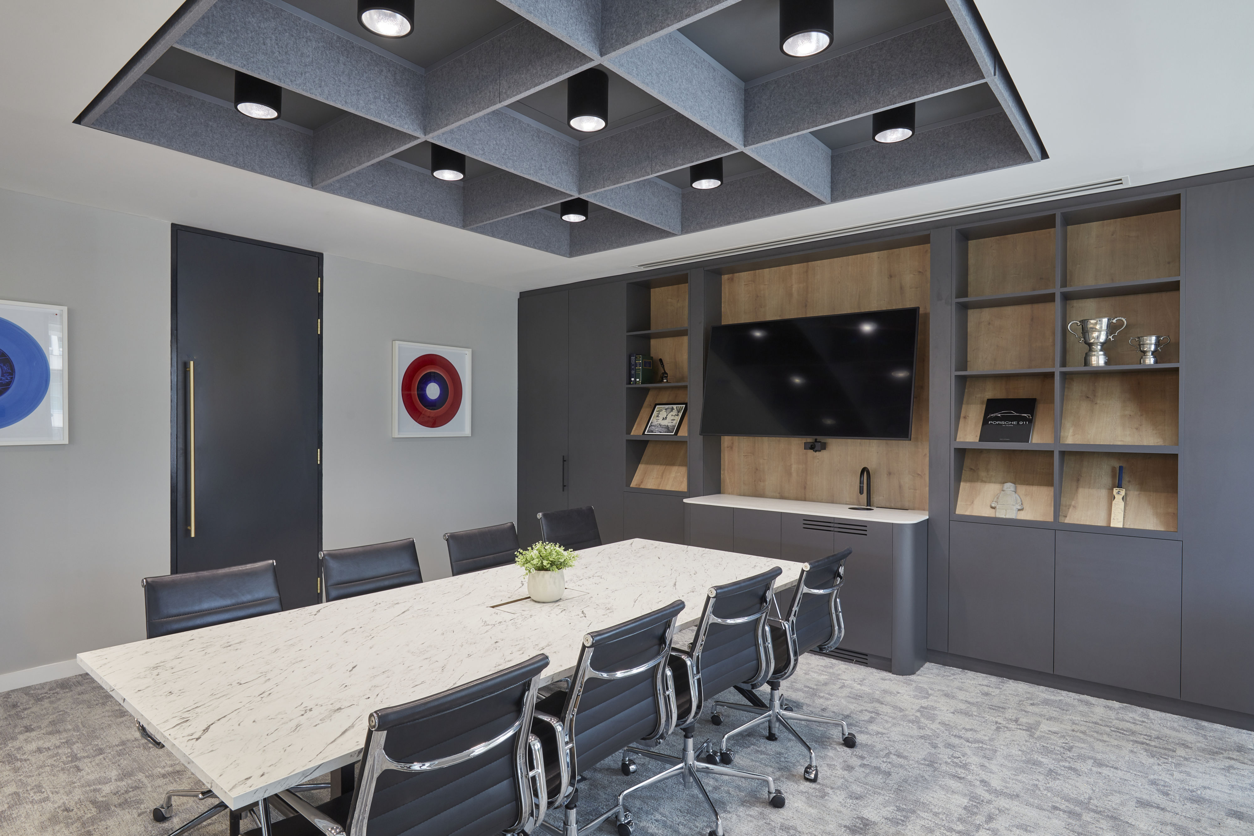 Conference room featuring a marble table with black chairs, a mounted television, and wall shelves holding trophies and books, under modern ceiling lights. Framed circular art decorates the walls.