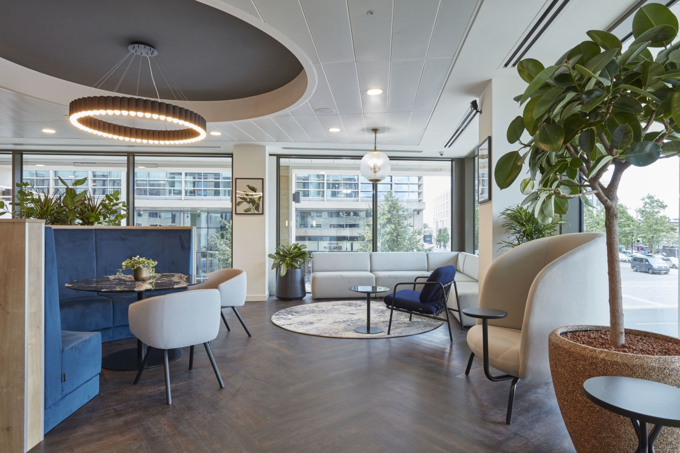 Modern office lounge with large windows, circular wood table, cushioned blue booth, and circular ceiling light. Green plants add a fresh touch