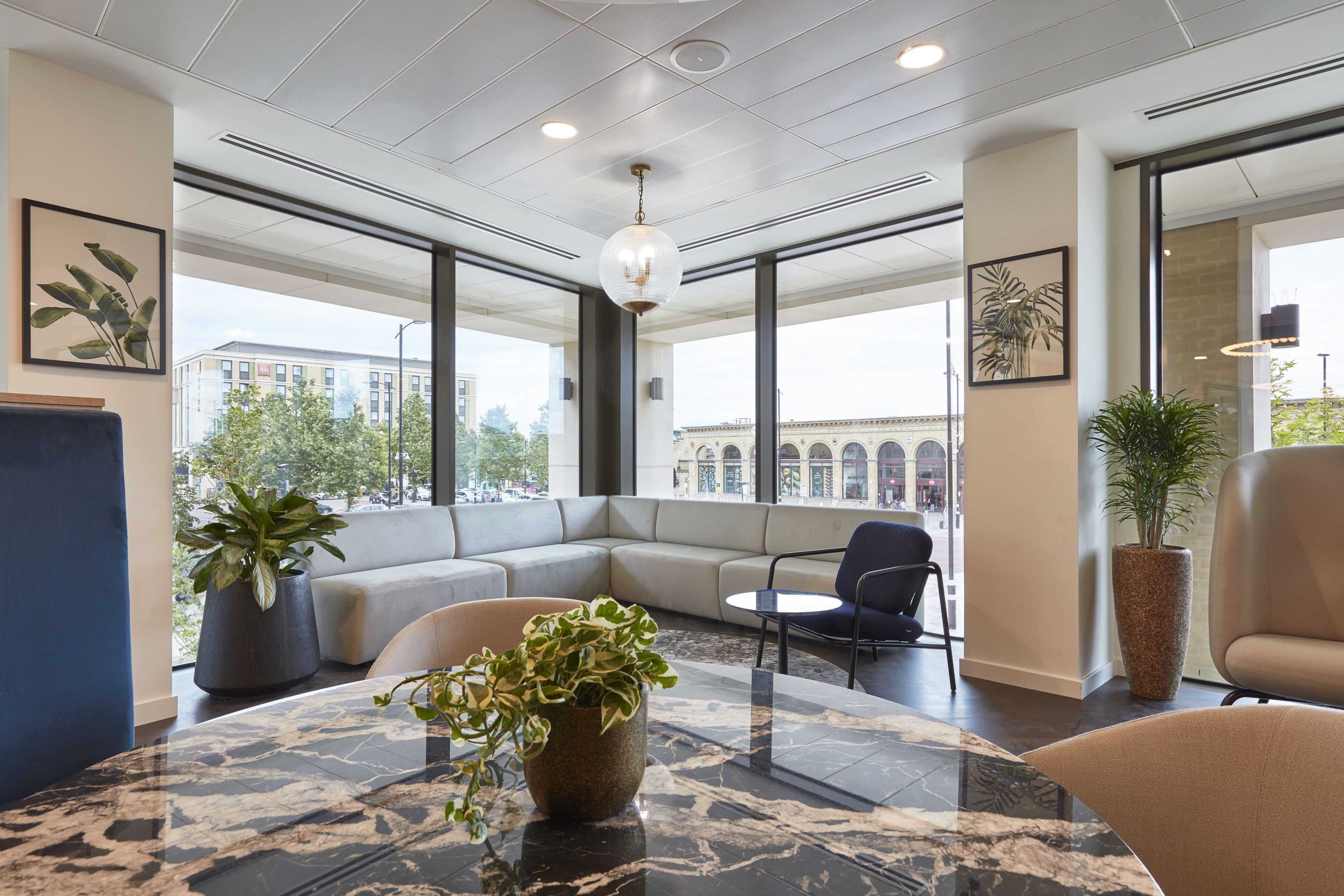 Corner lounge area with a grey L-shaped sofa and a round table, surrounded by large windows showcasing an urban street view, decorated with potted plants and framed botanical prints.