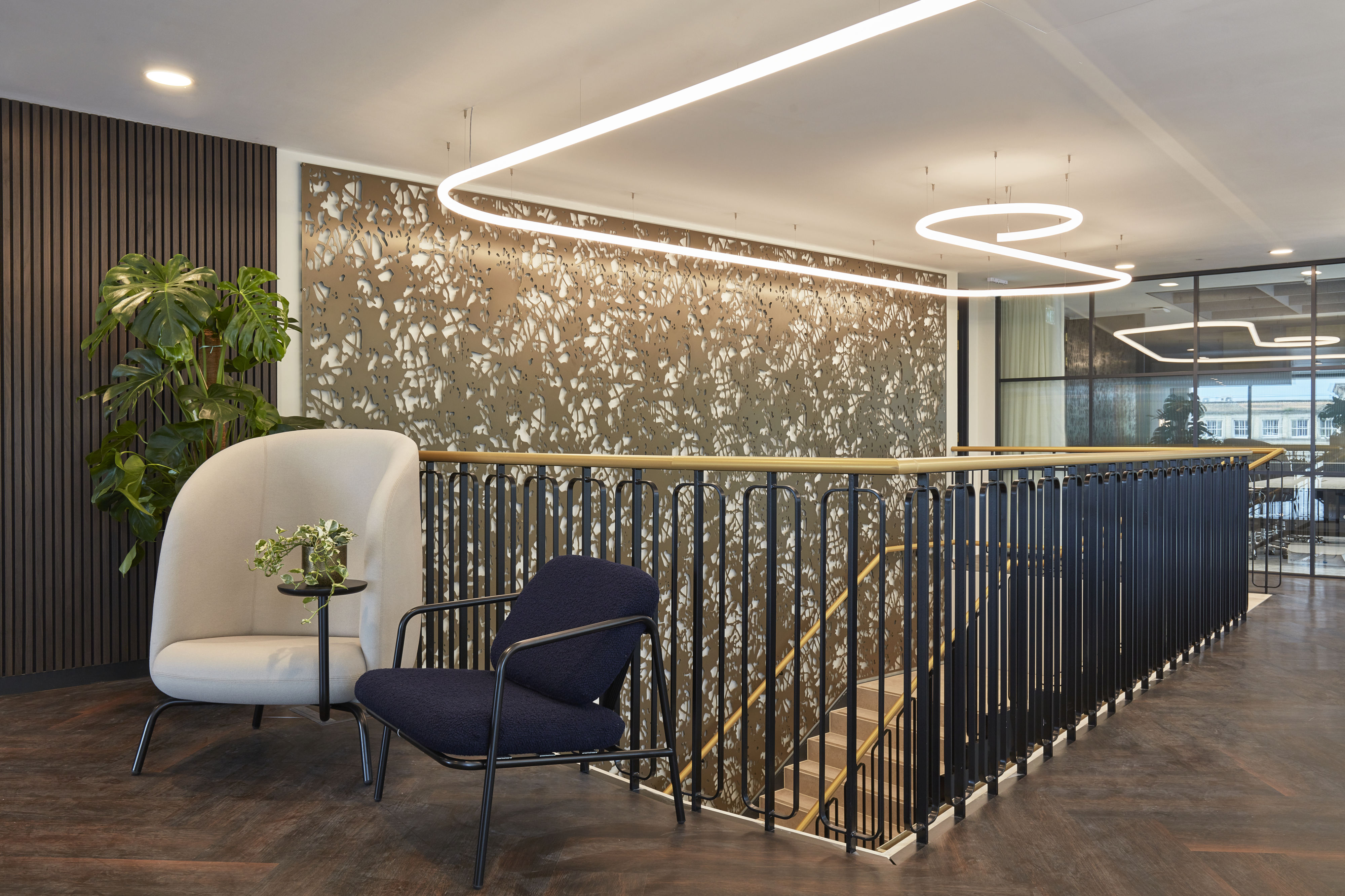 A modern office interior features two chairs and a small round table with a plant, near a staircase with ornate metal railing and overhead lighting, set against a patterned wall.
