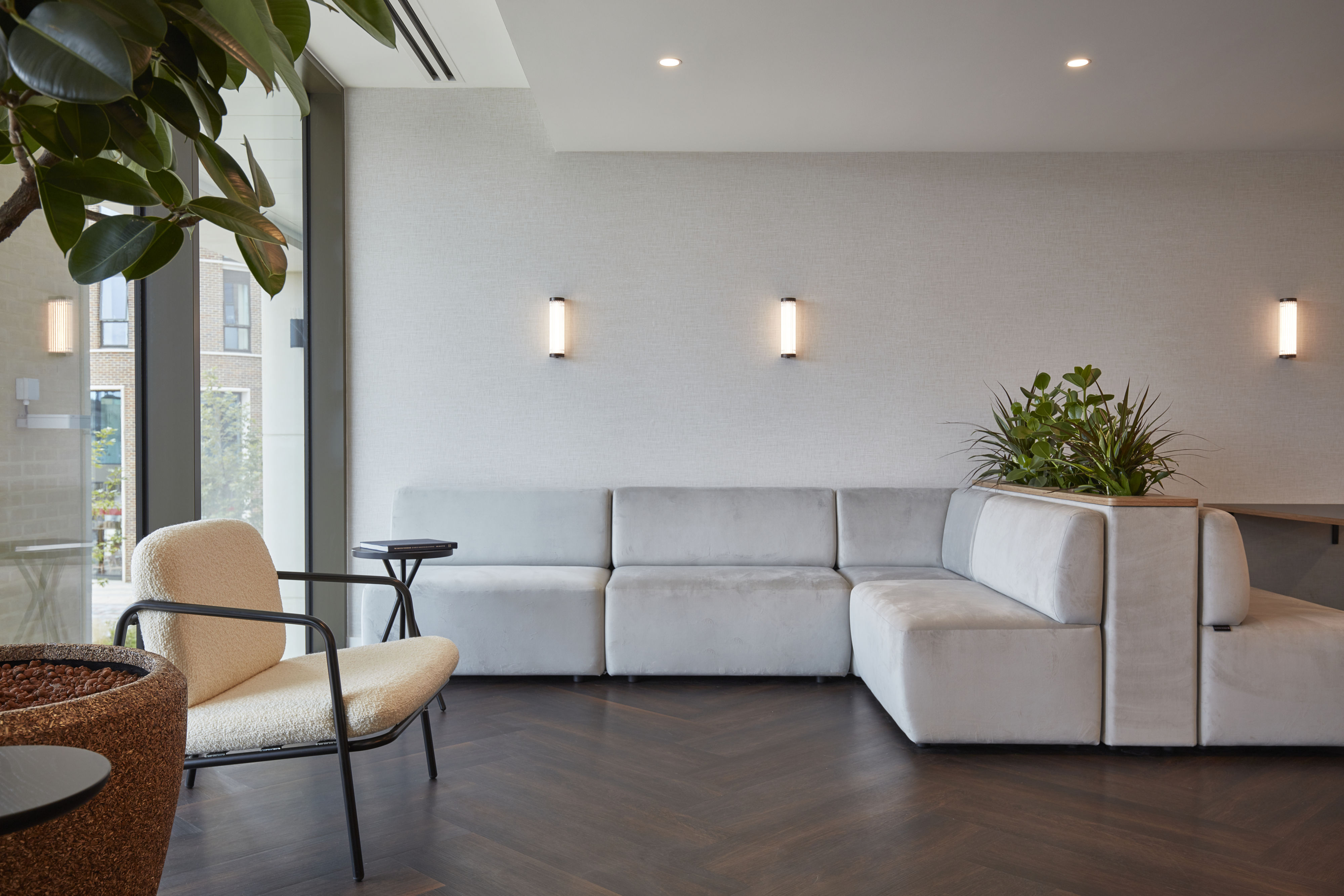A modern lounge features a gray sectional sofa, an upholstered armchair, and a small table with a book. Indoor plants enhance the room, illuminated by wall lights and natural sunlight.